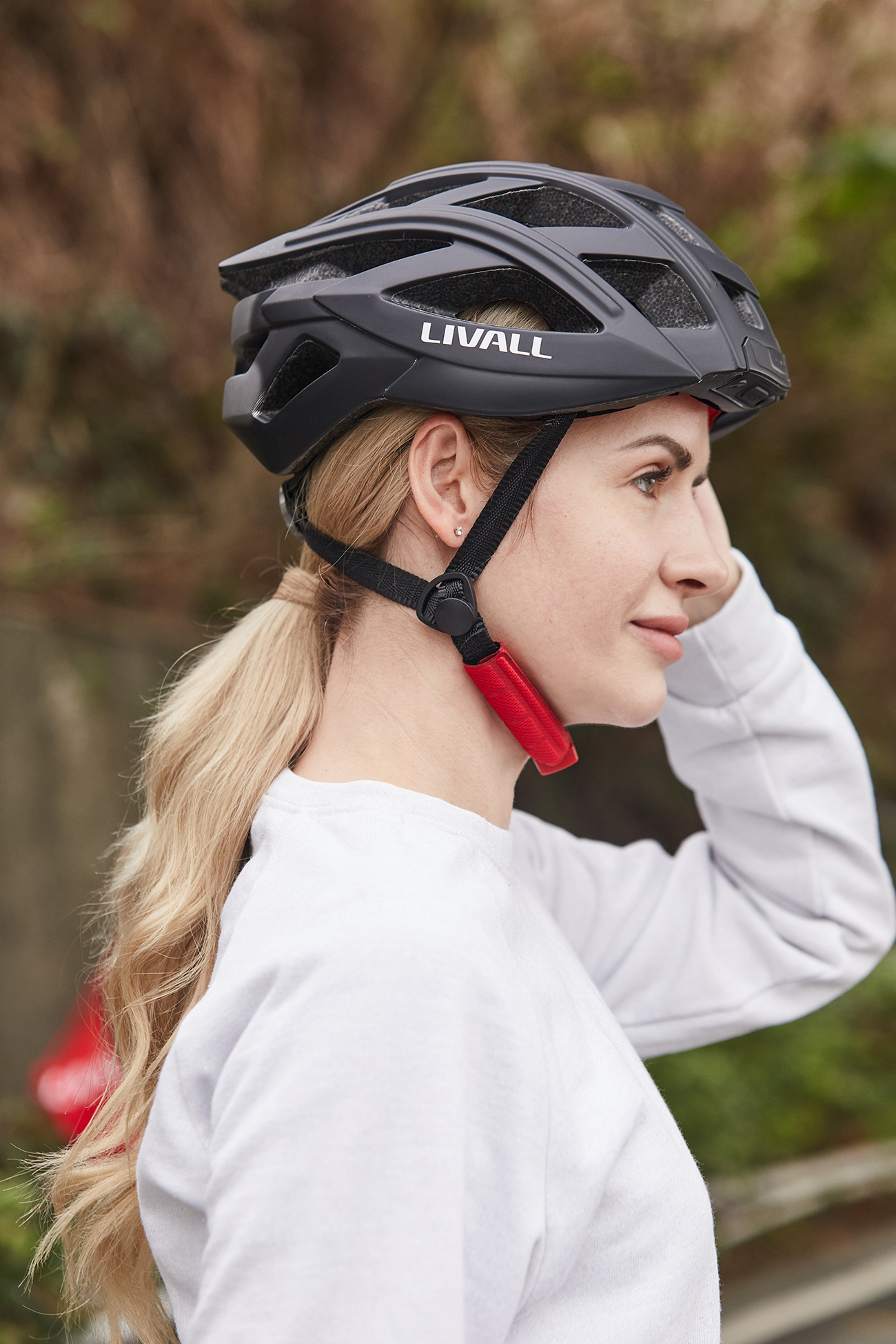 Eine Frau mit schwarzem LIVALL-Helm, blondem Haar und weißem Hemd. Der Hintergrund ist verschwommenes Grün.