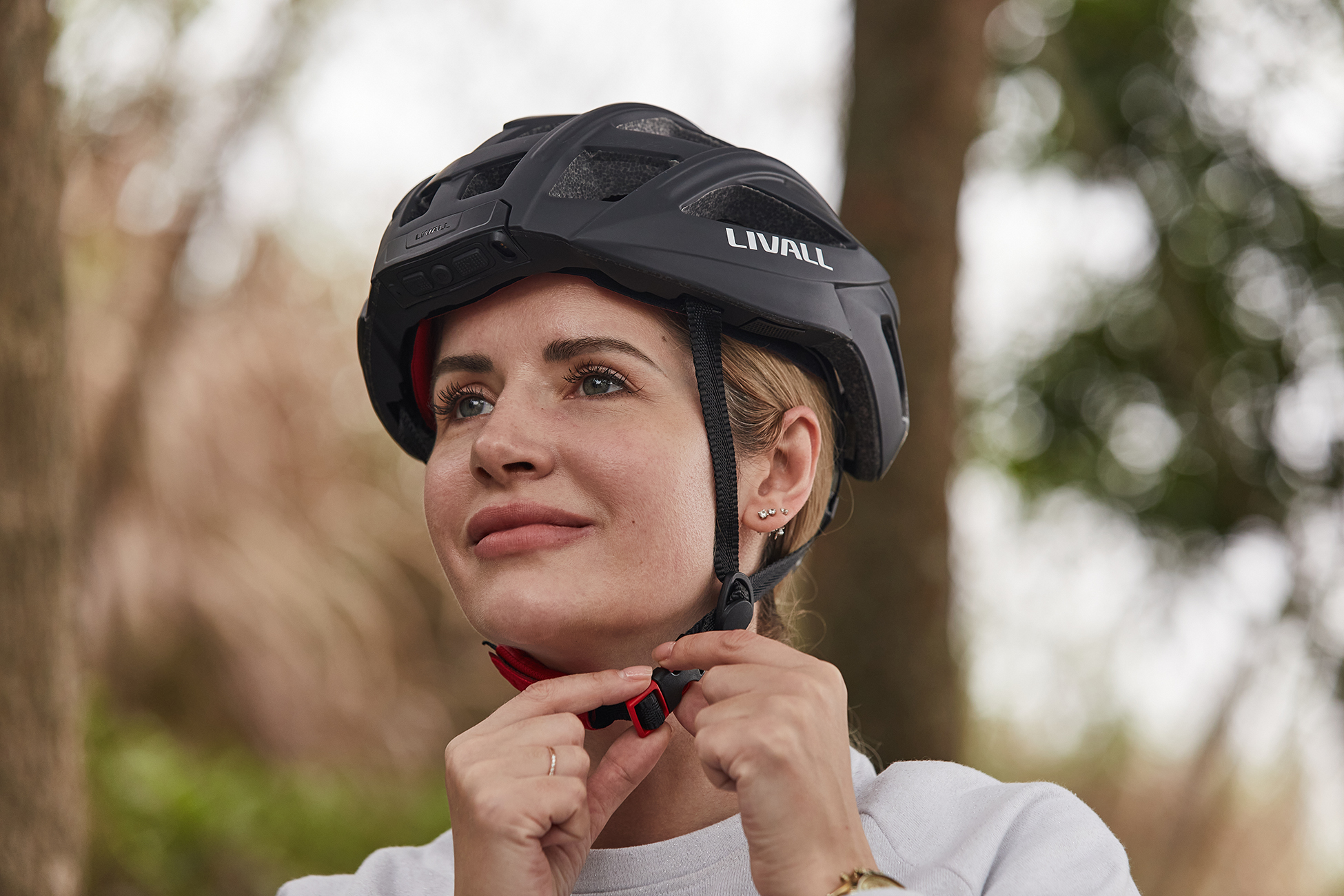Eine Frau mit schwarzem LIVALL-Helm befestigt den Kinnriemen. Sie ist hellhäutig mit blondem Haar. Bäume im Hintergrund.