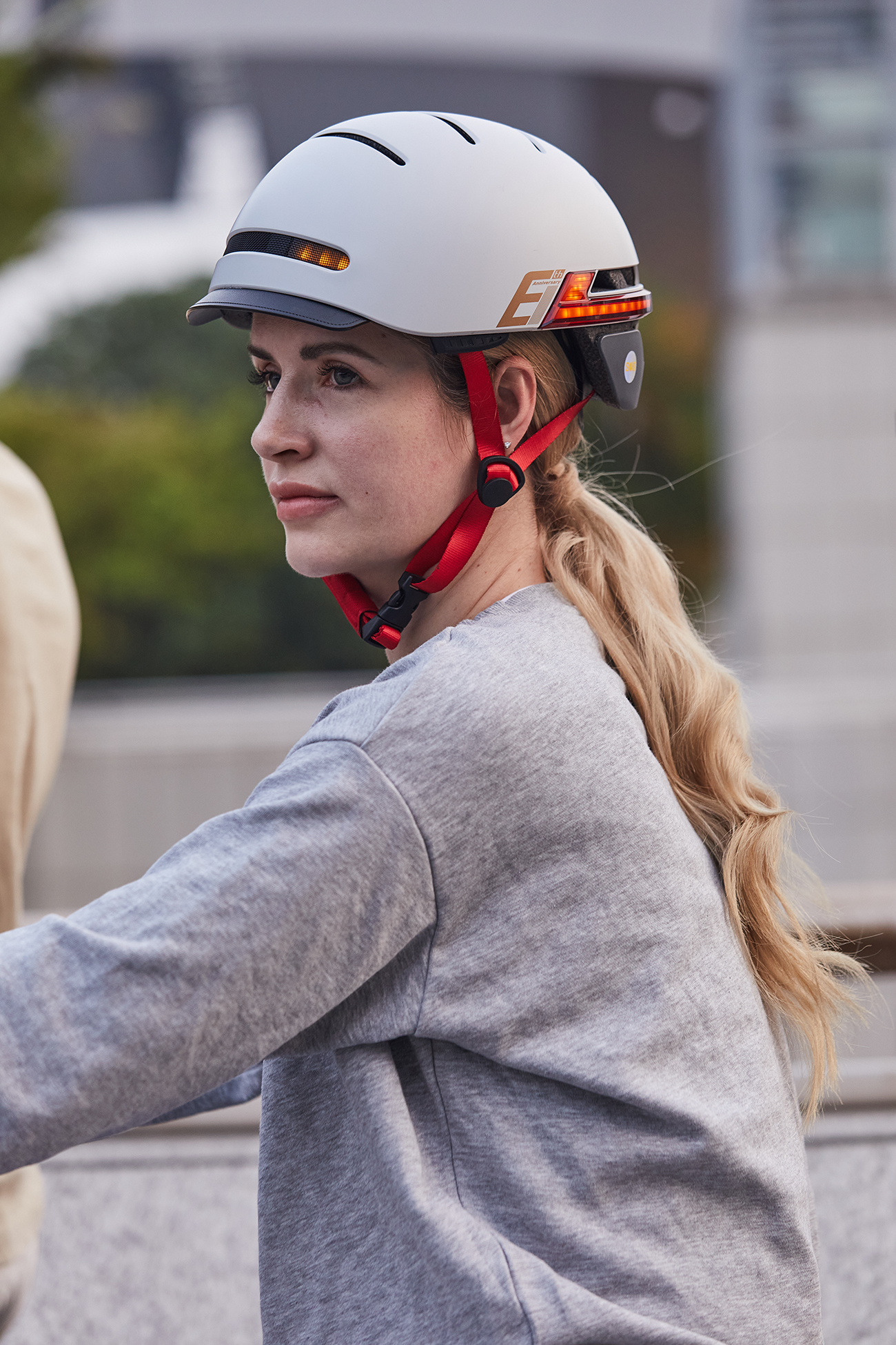 Eine Frau mit blonden Haaren trägt einen weißen Helm mit rotem Riemen. Sie trägt einen grauen Pullover. Der Hintergrund ist unscharf.