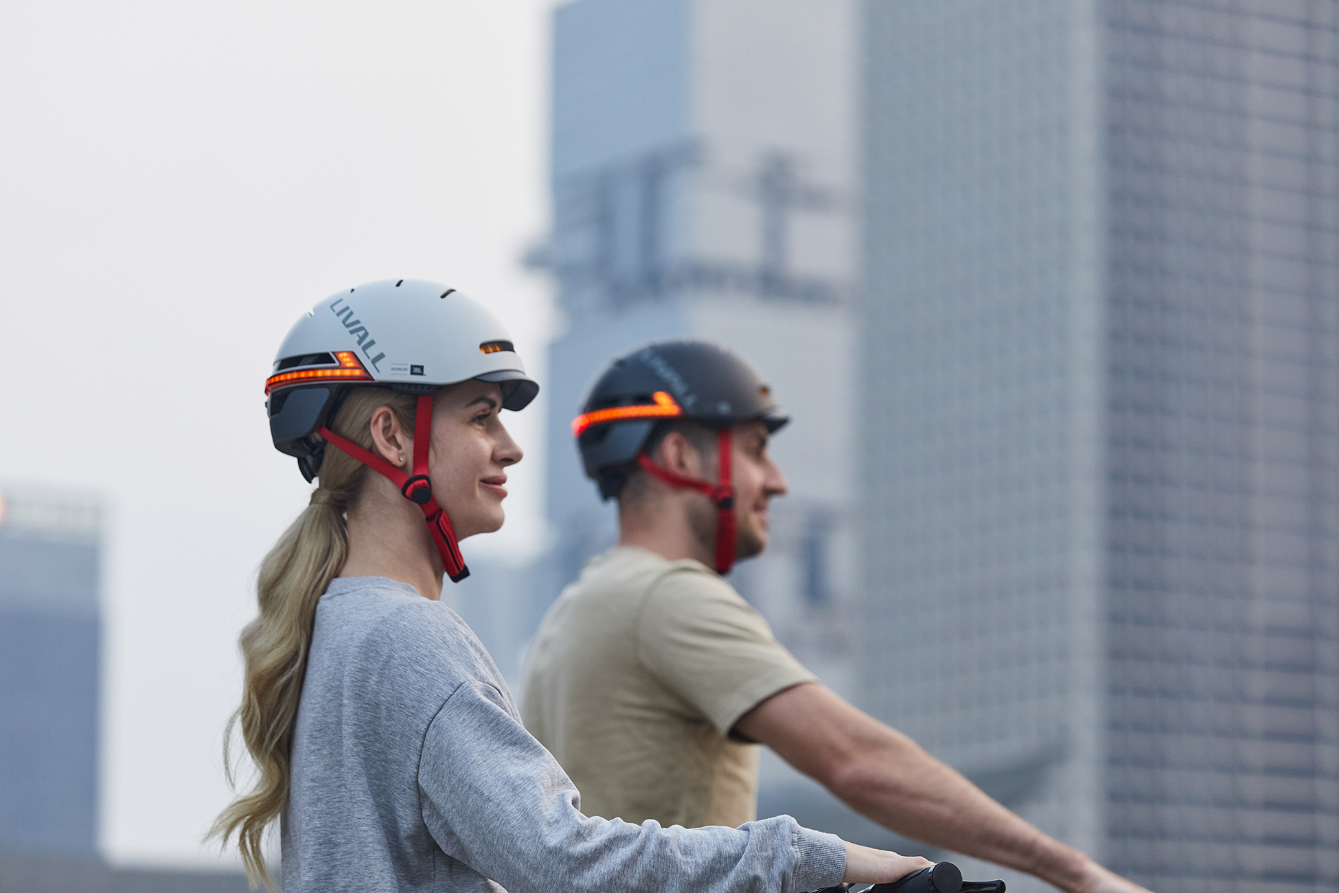 Zwei Personen tragen Helme mit Lichtern und fahren Fahrräder. Gebäude sind im Hintergrund unscharf. Der Himmel ist leicht bewölkt.