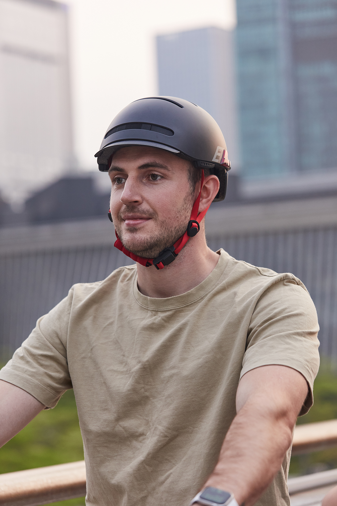 Mann mit schwarzem Helm und roten Riemen, beigem Hemd, lächelnd, mit Stadtgebäuden im Hintergrund.