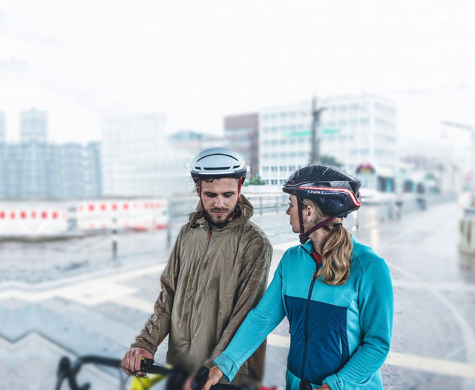 Due persone con caschi e giacche in piedi accanto a una bicicletta. Edifici della città sullo sfondo sfocato. Sembra che stia piovendo.