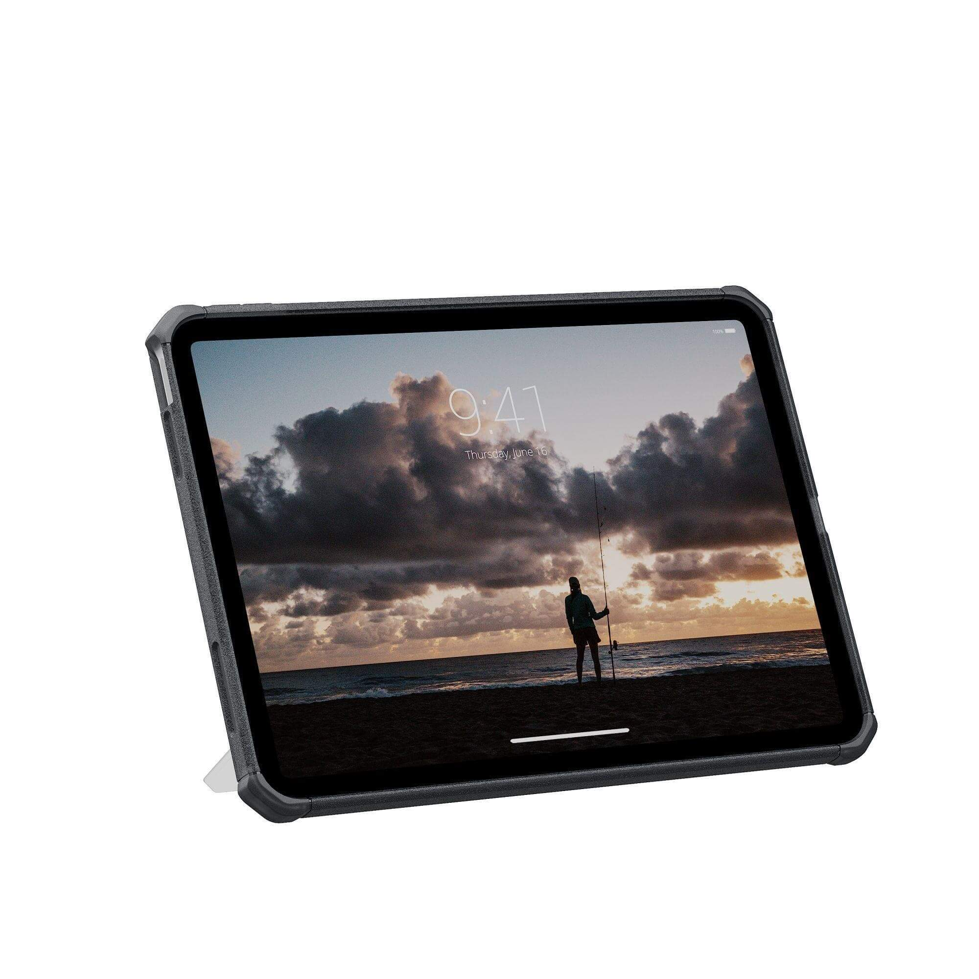 Tablet wyświetlający scenę plażową z osobą łowiącą ryby. Ciemne etui, biała podstawka. Pochmurne niebo i zachód słońca.
