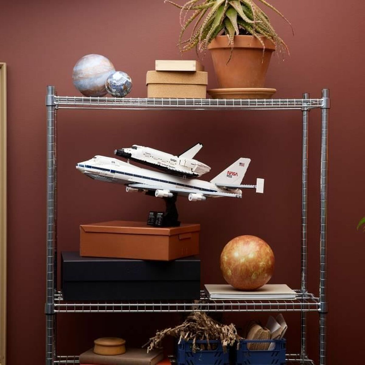 Metalowa półka z modelem wahadłowca kosmicznego na samolocie. Inne elementy to pudełka, roślina i kule. Tło jest ciemnoczerwone.