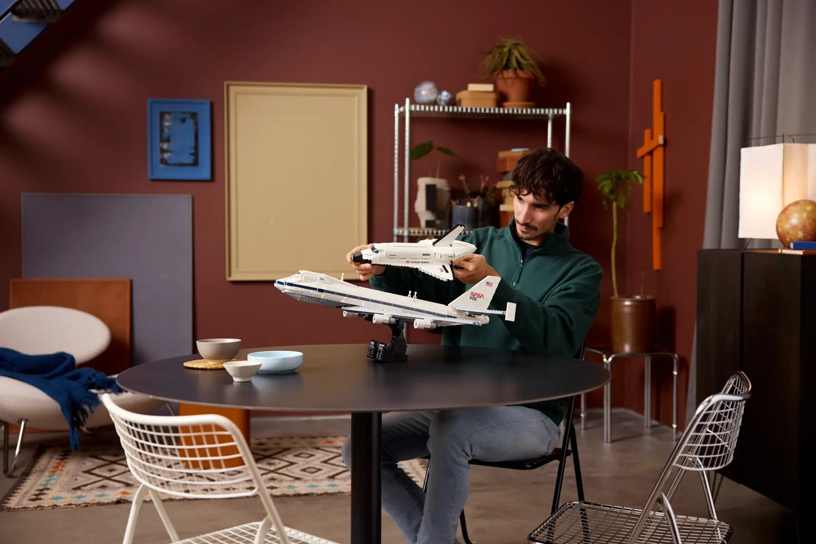 Mężczyzna trzyma biały model samolotu NASA i wahadłowiec kosmiczny. Siedzi przy okrągłym stole z kilkoma miseczkami i krzesłami.