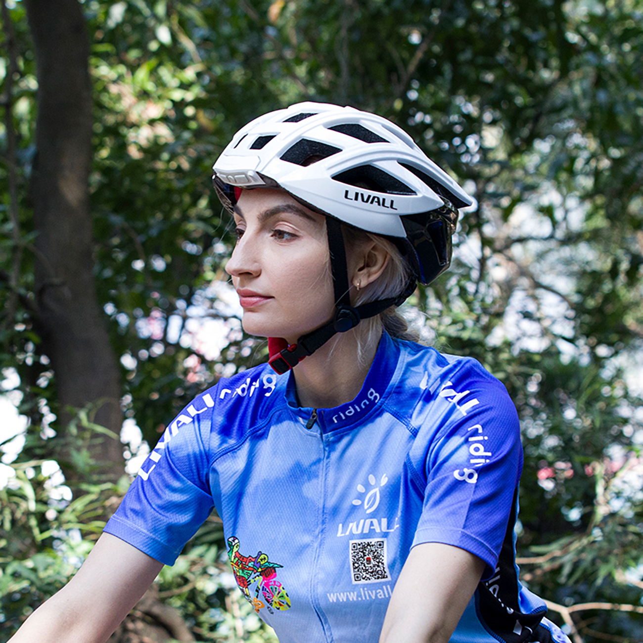 Eine Frau mit weißem Helm und blauem Radtrikot blickt zur Seite. Bäume bilden den Hintergrund. Auf dem Helm steht LIVALL.