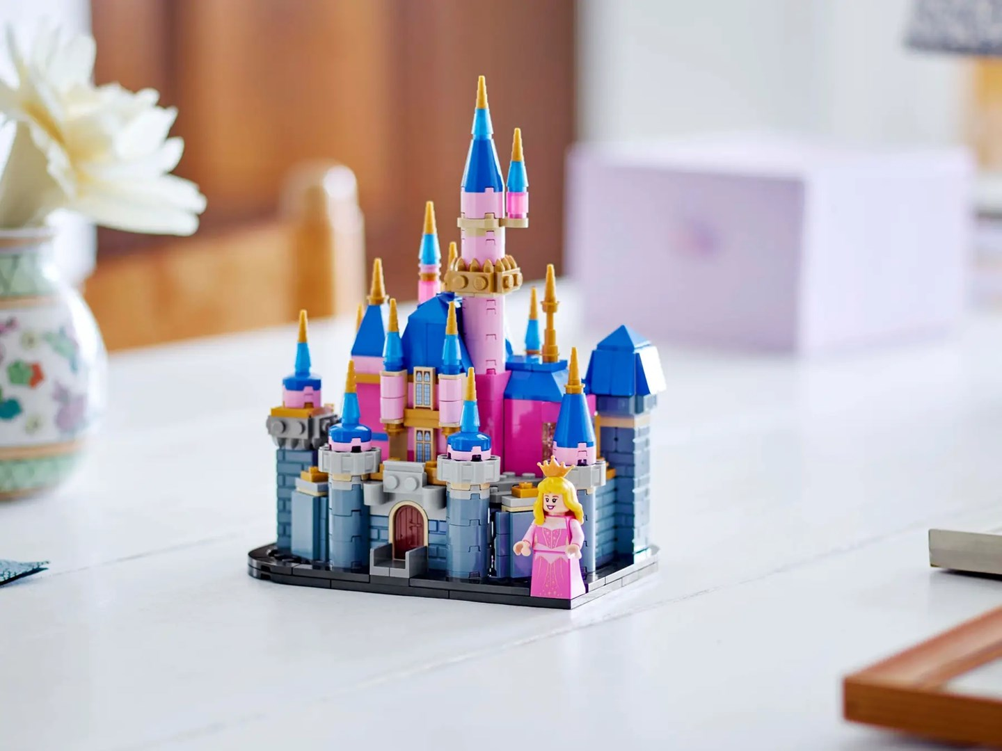 Zamek Lego Disney z niebieskimi, różowymi i szarymi klockami. Figurka księżniczki stoi z przodu. W tle wazon i pudełko.