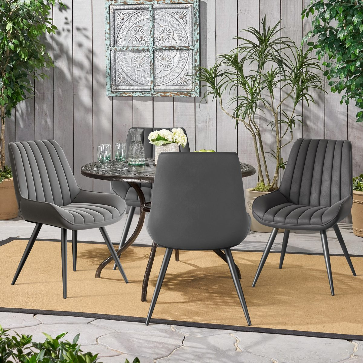 Essgruppe im Freien: Tisch, vier graue Stühle, Teppich, Pflanzen und Wanddekoration. Die Szene spielt auf einer Terrasse mit einer verwitterten Wand.
