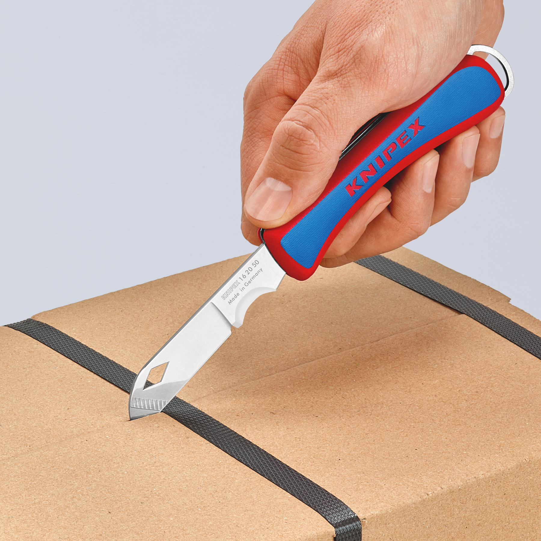 Una mano sosteniendo un cuchillo rojo y azul cortando una caja de cartón. La hoja del cuchillo es plateada.