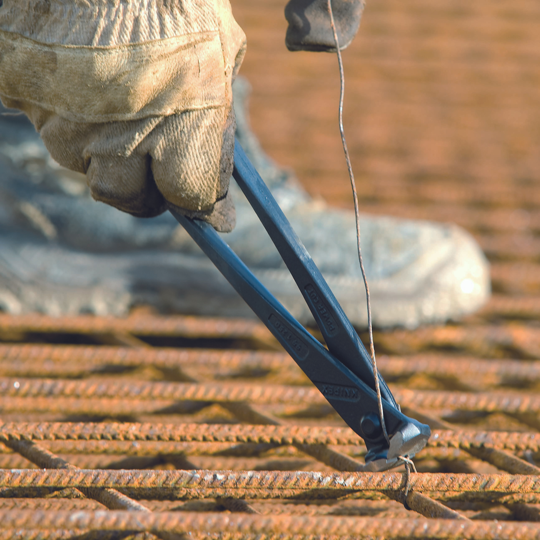Eine behandschuhte Hand verdreht mit einer Zange Draht auf einem Metallgitter. Ein Stiefel ist im Hintergrund zu sehen.