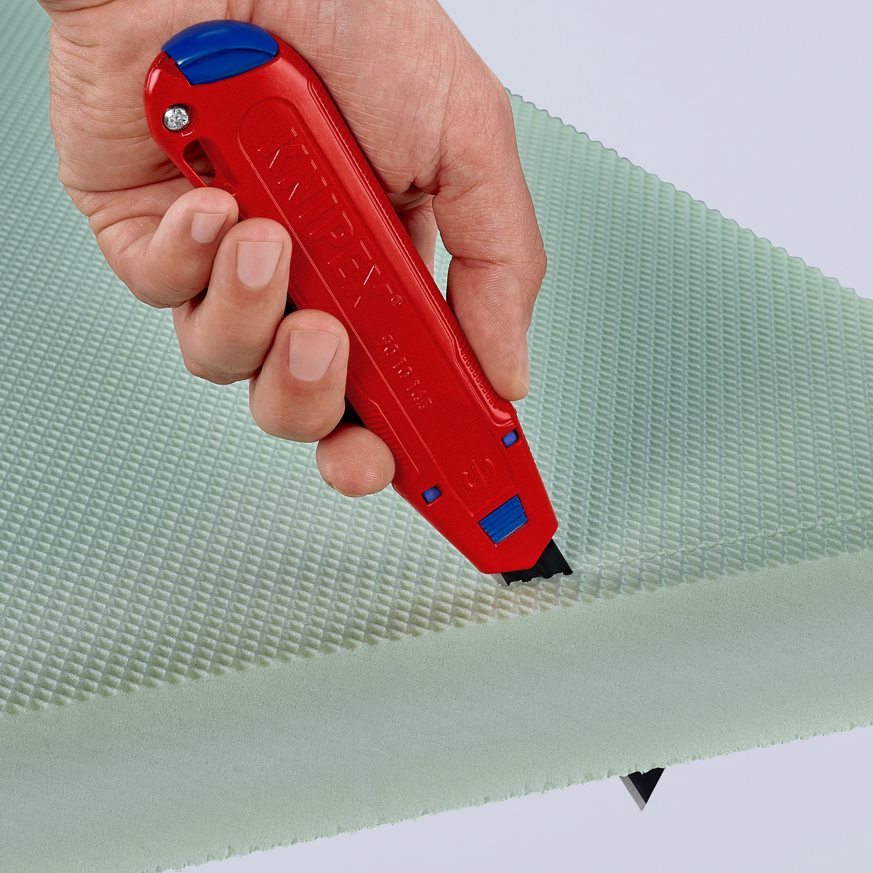 Eine Hand hält ein rotes Universalmesser, das einen hellgrünen Schaumstoffblock schneidet. Das Messer ist schwarz und blau.