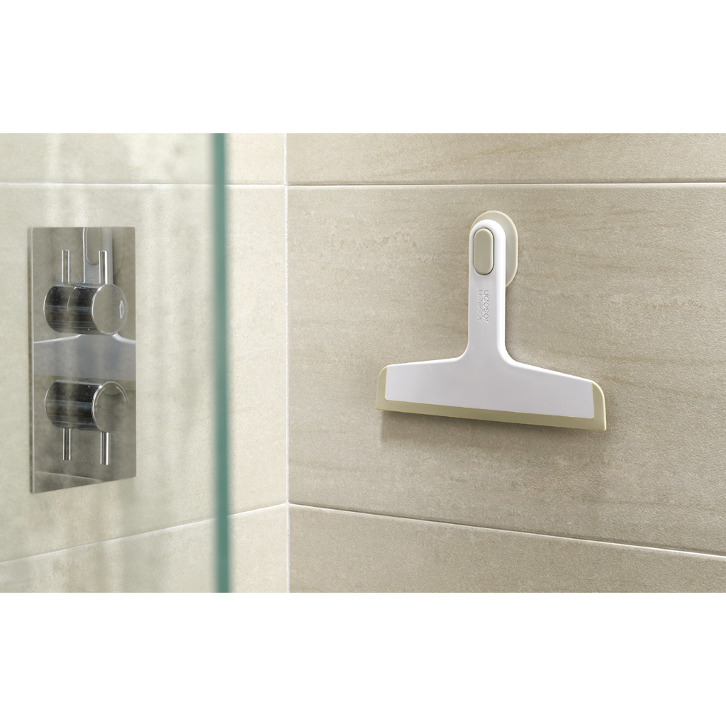 Un limpiacristales blanco con una hoja gris está montado en una pared alicatada. A la izquierda se ve un mando de ducha cromado.