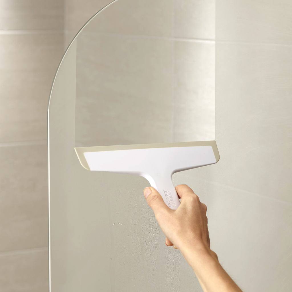 Una mano sostiene un limpiacristales blanco en una puerta de ducha de cristal. El fondo muestra azulejos beige.