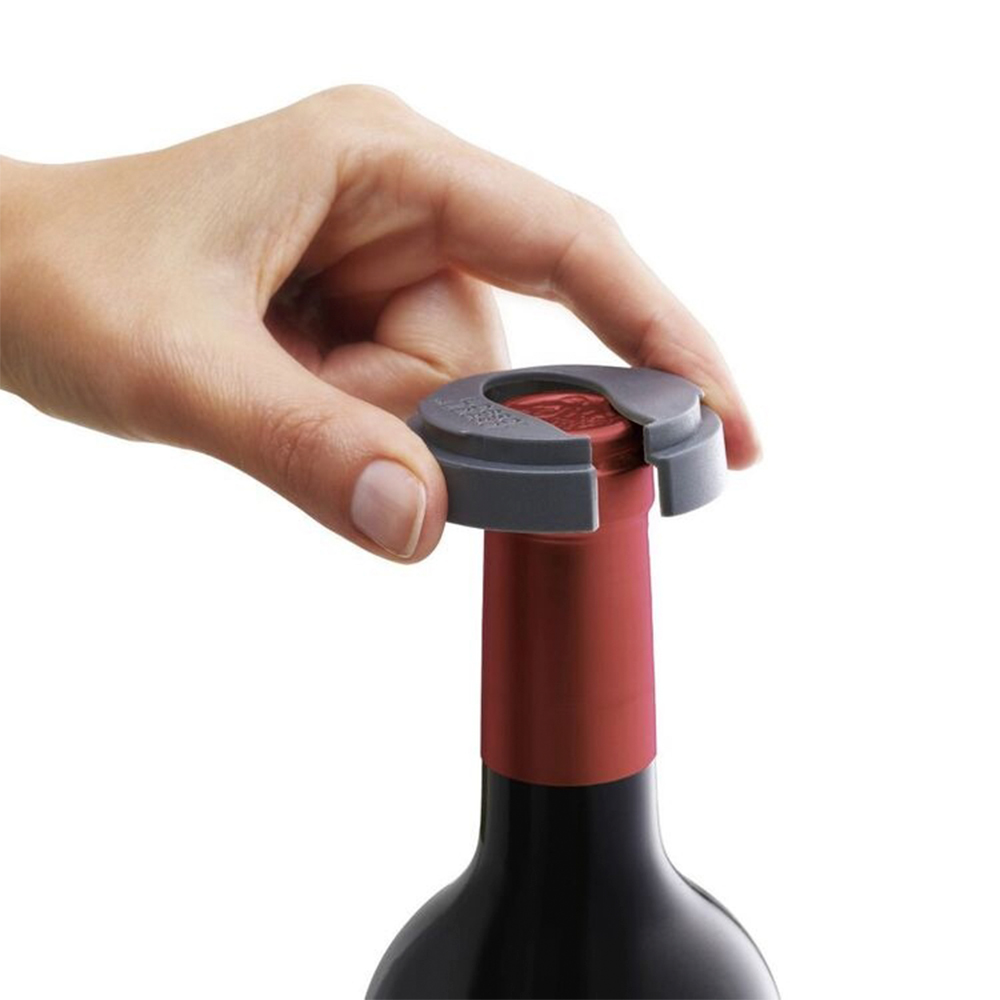 Una mano usa un abridor de botellas de vino gris para quitar el sello rojo de una botella de vino. La botella es negra y roja.