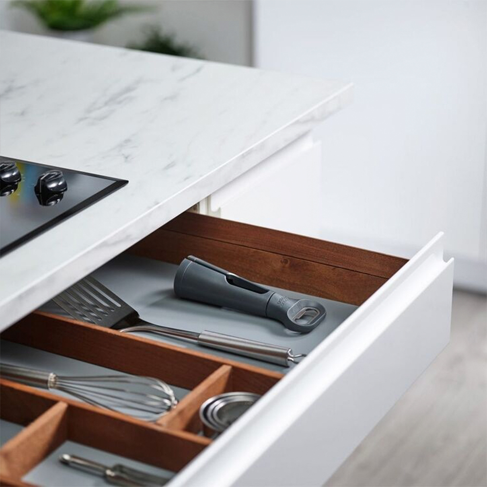 Cajón de cocina abierto con utensilios: espátula, batidor, abridor de botellas y más. Encimera blanca y divisores de cajones de madera visibles.