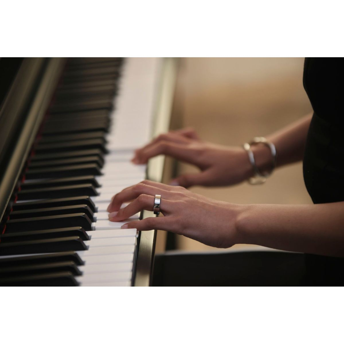 Hände spielen Klavier. Nahaufnahme zeigt Hände auf Tasten. Ein Ring ist sichtbar. Ein Armband ist am Handgelenk. Schwarzes Klavier.