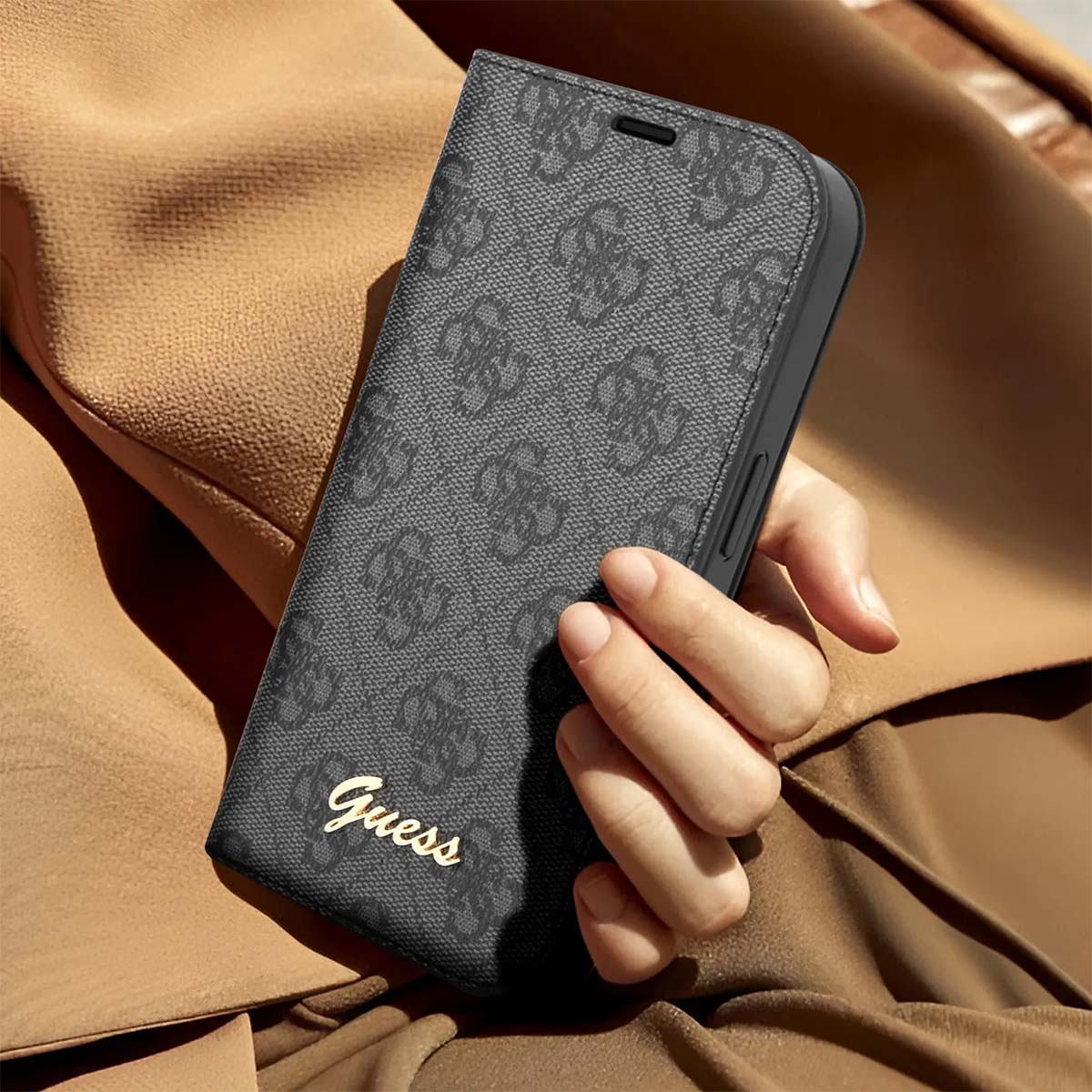Una persona tiene in mano una custodia per telefono grigio scuro con un motivo ripetuto e il logo Guess. Lo sfondo è una giacca beige.