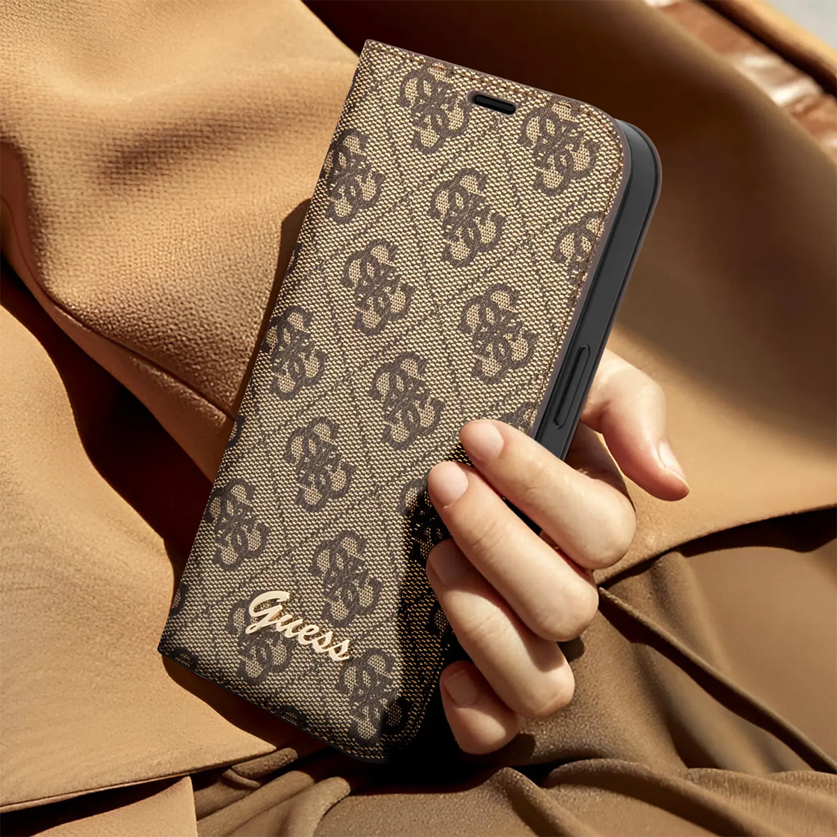Una mano tiene una custodia per telefono Guess. La custodia è marrone con un motivo a logo ripetuto. Lo sfondo è una giacca marrone.