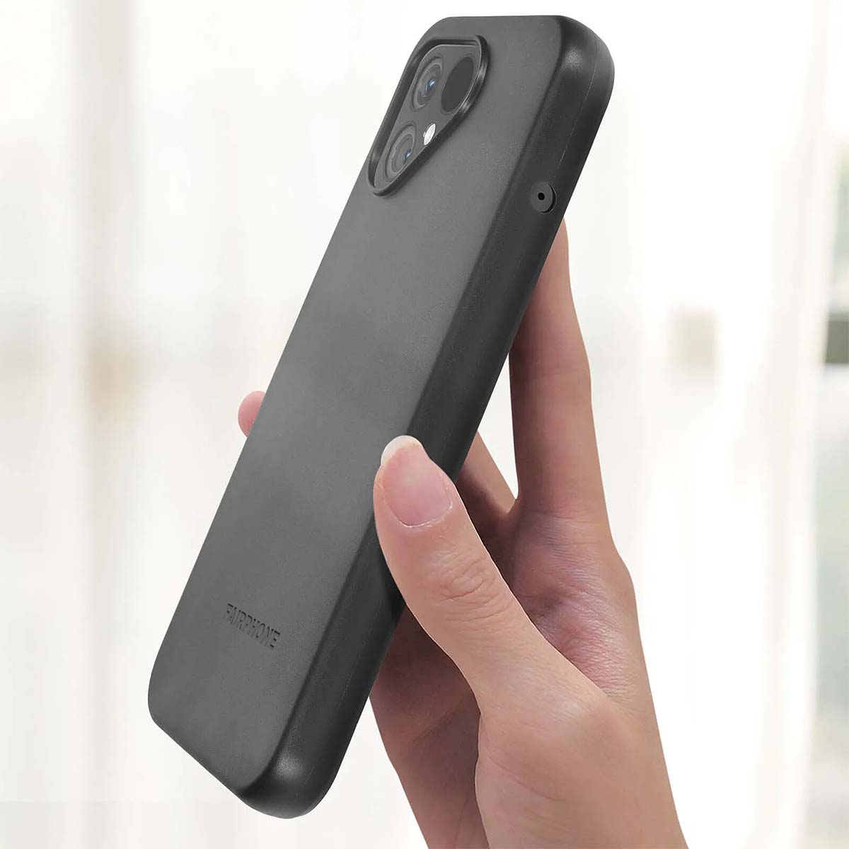 Una mano che tiene una custodia nera per telefono. La custodia ha un ritaglio per la fotocamera e la scritta "FAIRPHONE". Lo sfondo è sfocato.