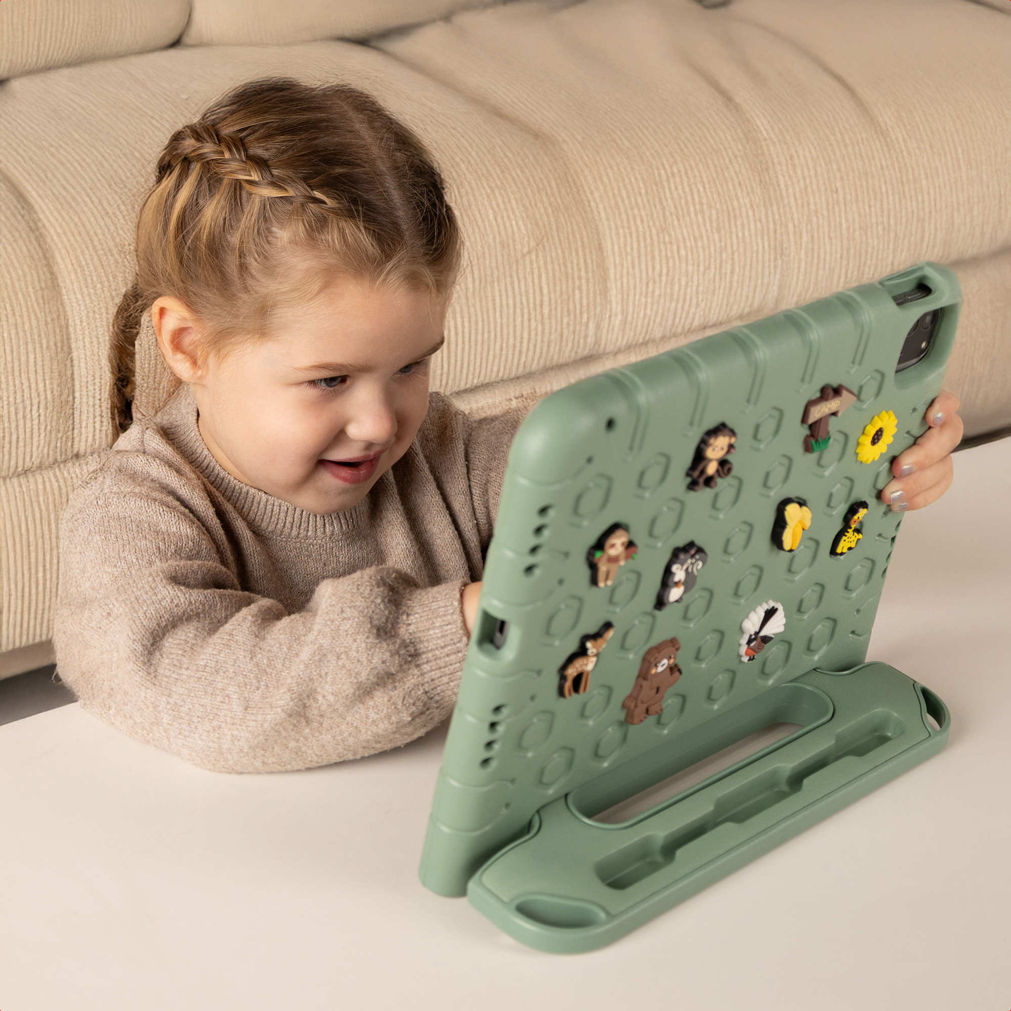 Een meisje met een vlecht bekijkt een tablet met stickers. De tablet staat op een witte tafel. Op de achtergrond is een beige bank.