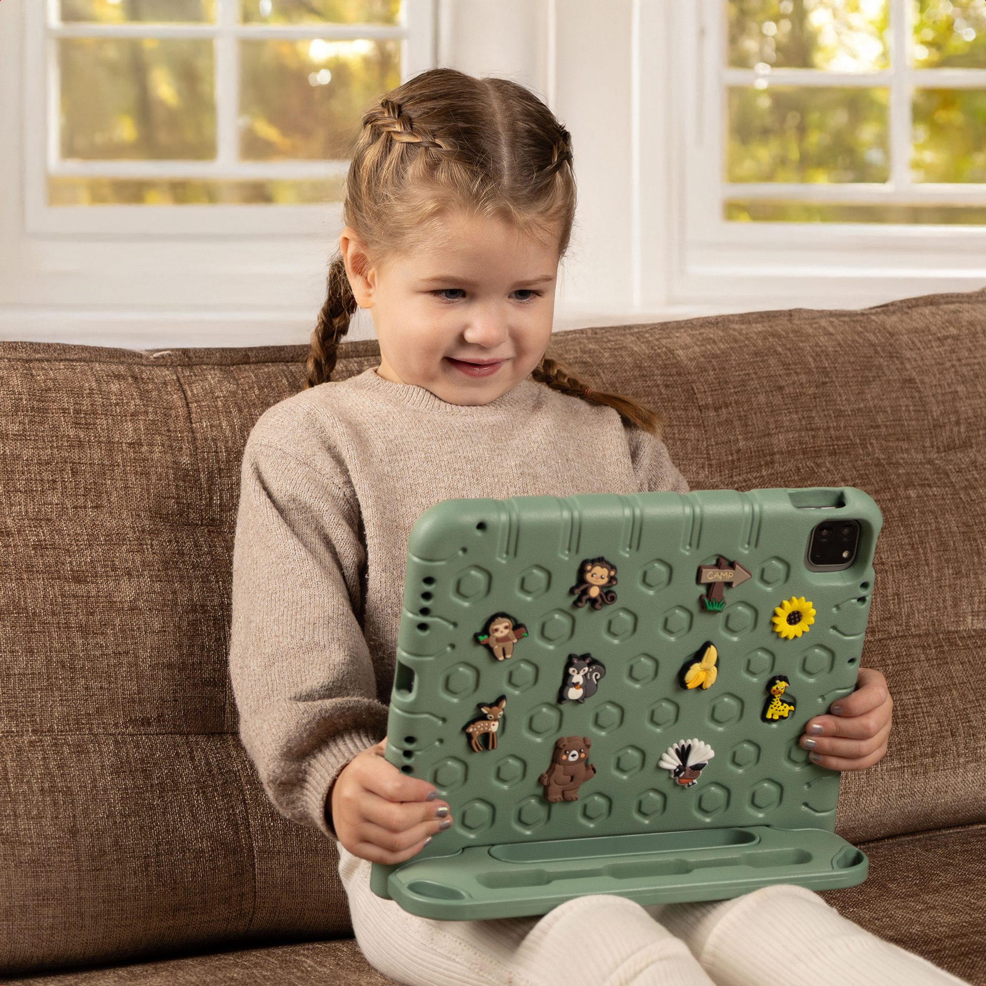Een meisje met vlechten houdt een groene tablet vast, versierd met dierenstickers, zittend op een bruine bank. Achtergrond is een raam.