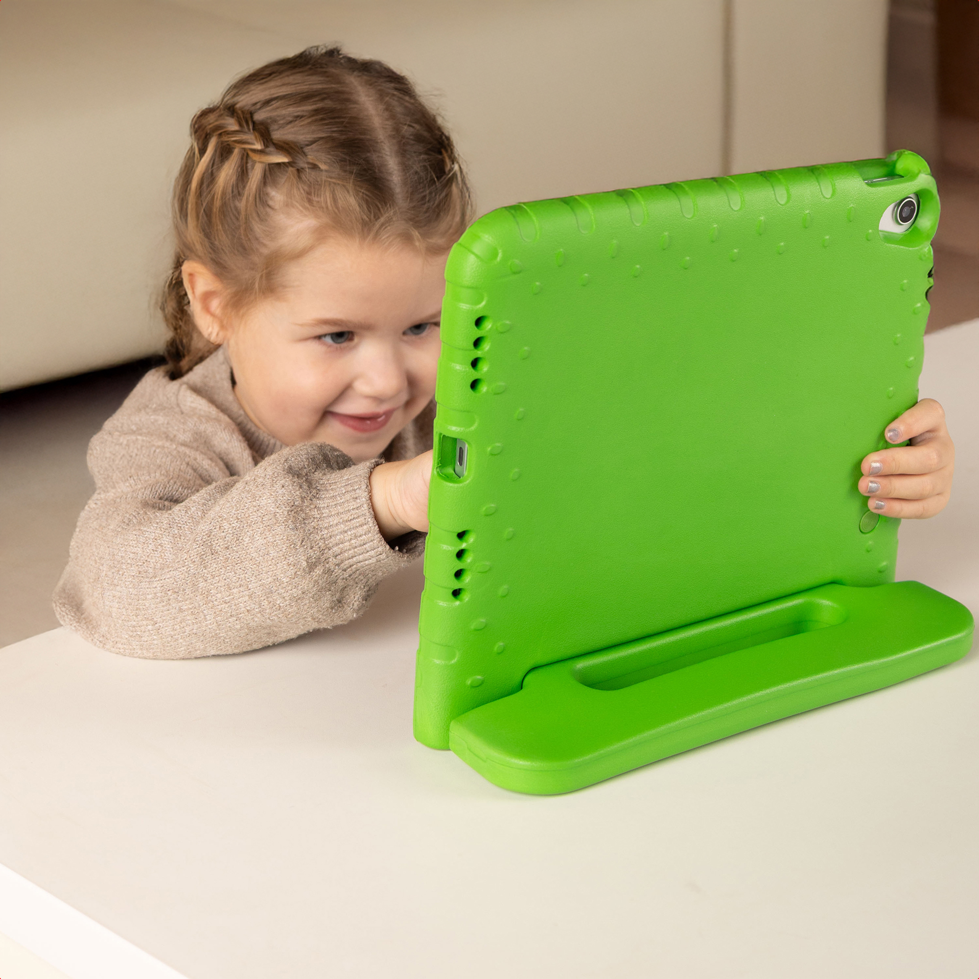 Een jong meisje lacht naar een groene tablet. Ze draagt een beige trui en de tablet staat op een wit oppervlak.