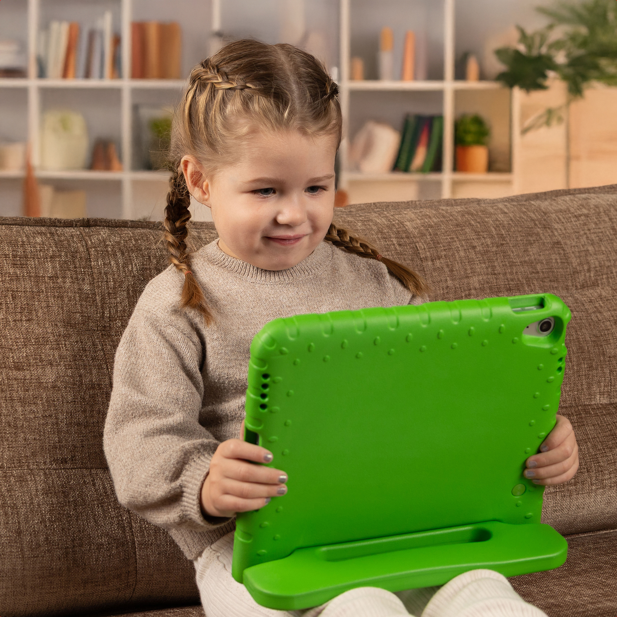 Een meisje met vlechten zit op een bank en houdt een groene tablet vast. Achtergrond heeft een boekenplank en planten.