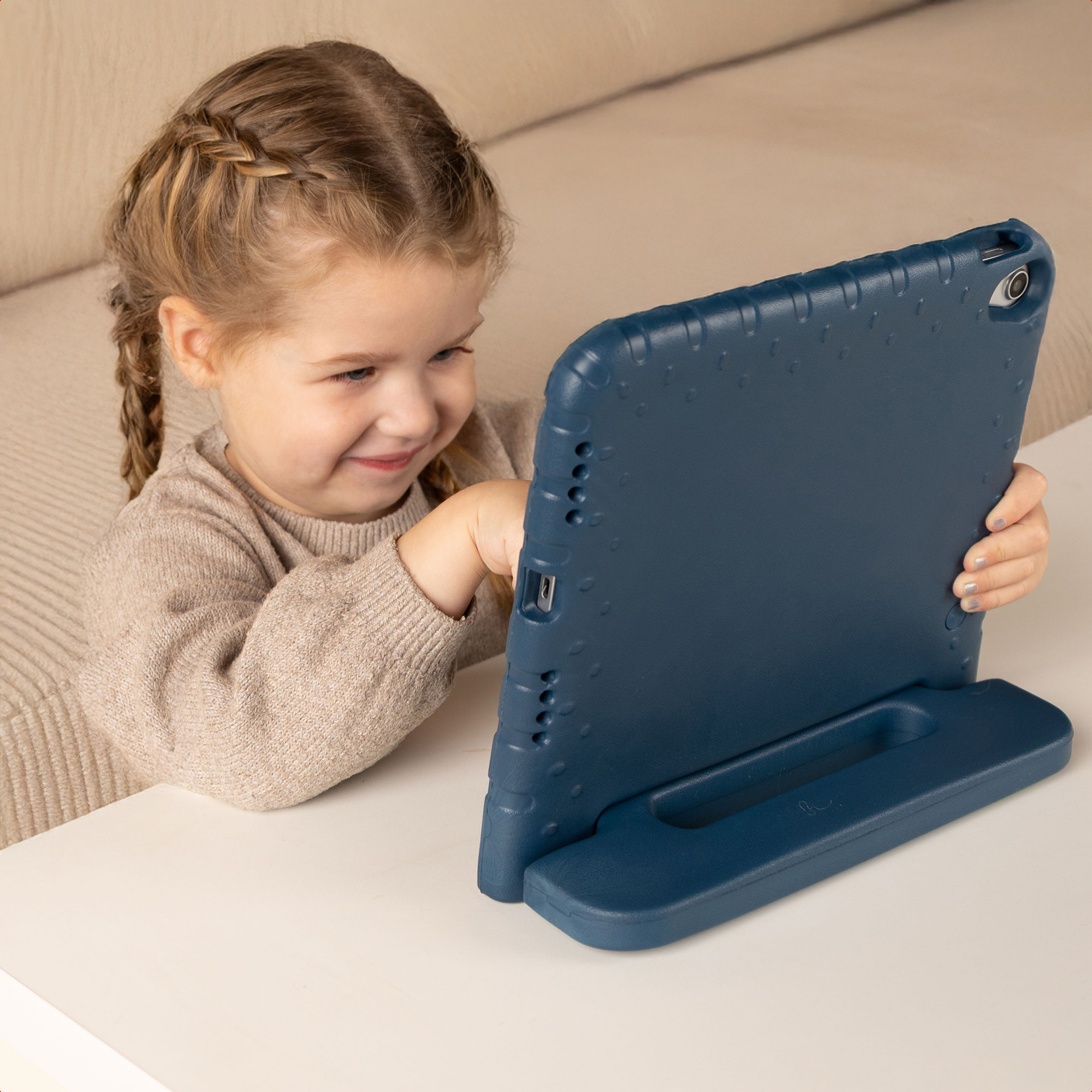 Een lachend meisje met vlechten communiceert met een blauwe tablet op een witte tafel. Beige achtergrond met een bank.