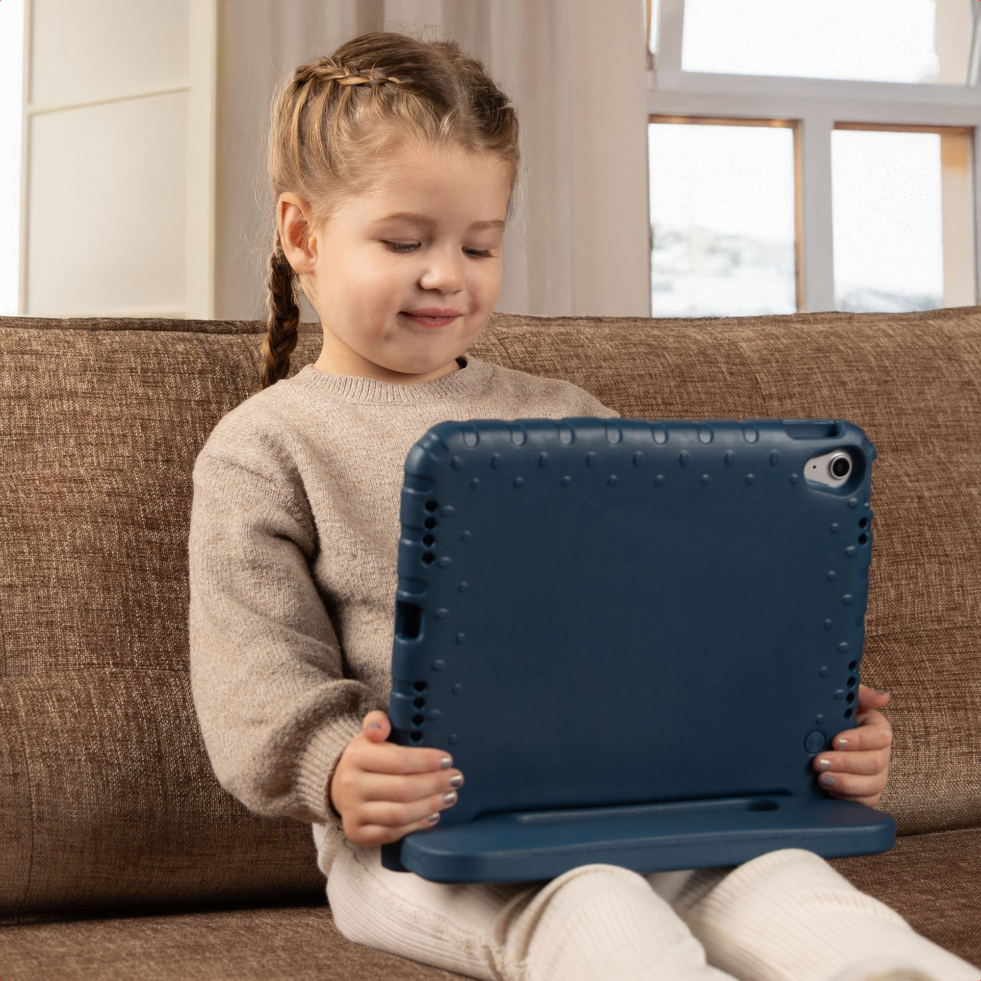 Een meisje met vlechten zit op een bank en houdt een blauwe tablet vast. Ze glimlacht terwijl ze naar het scherm kijkt. De achtergrond is een raam.