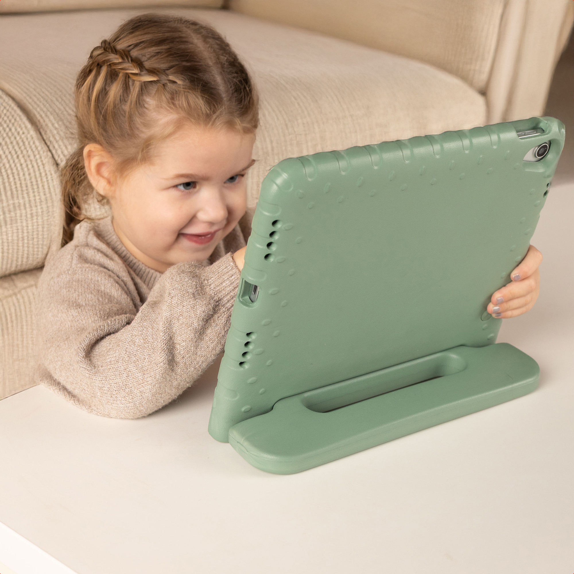 Een jong meisje met blond haar glimlacht terwijl ze naar een groene tablet kijkt. De tablet staat op een witte tafel.