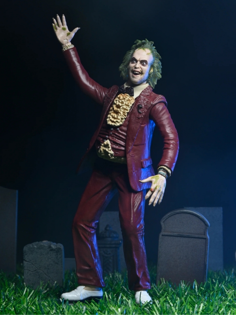Kırmızı takım elbiseli, yeşil saçlı ve beyaz ayakkabılı Beetlejuice figürü, mezar taşlarının önünde çimenlerin üzerinde duruyor.