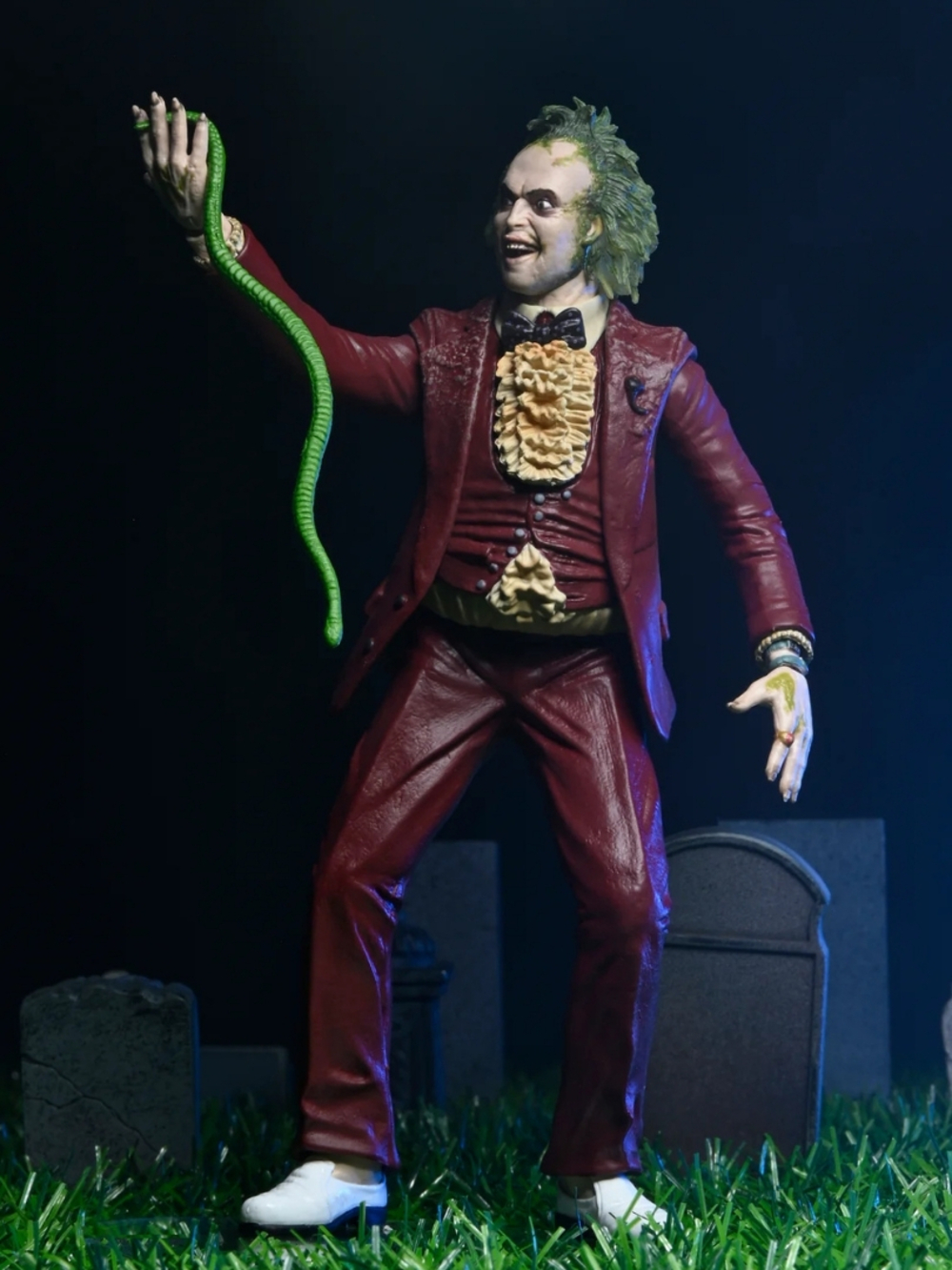 Bordo takım elbiseli Beetlejuice figürü, yeşil bir yılan tutuyor. Arka plan mezarlık temalı, mezar taşları ve yeşil çimenlerle.