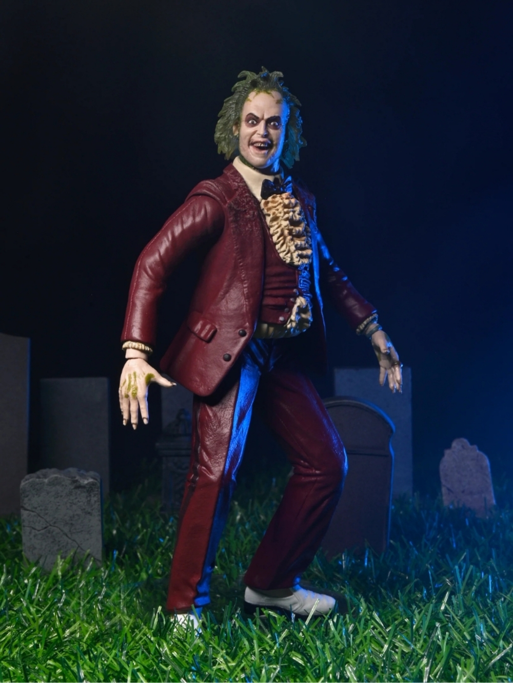 Burgonya renk takım elbiseli, beyaz ayakkabılı ve yeşil saçlı Beetlejuice figürü, yeşil çimenli bir mezarlıkta duruyor.