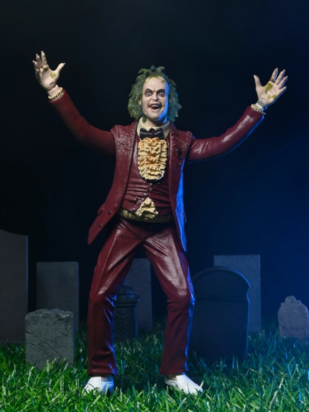 Beetlejuice figürü, mezarlıkta kolları havada duruyor. Bordo takım elbise, fırfırlı gömlek, yeşil saç ve beyaz ayakkabılar giyiyor.