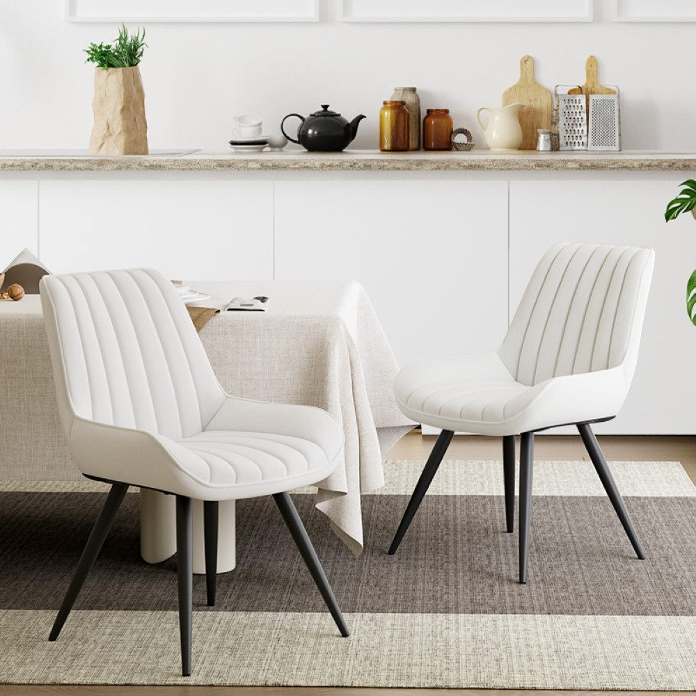 Zwei weiße Stühle mit schwarzen Beinen in einer Küche. Tisch mit beiger Tischdecke, Küchenutensilien im Hintergrund.