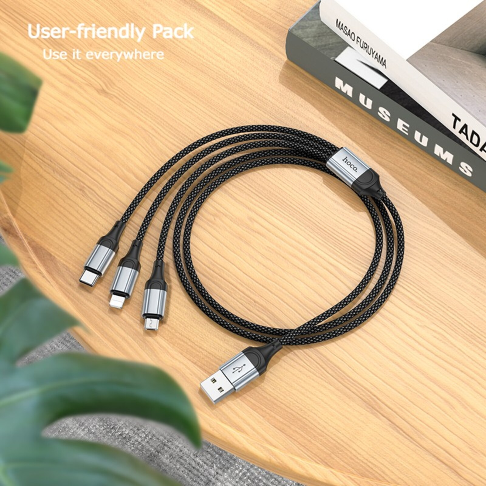Ahşap bir masa üzerinde, bir kitap ve bir bitkinin yanında, çoklu konektörlü siyah USB şarj kabloları.