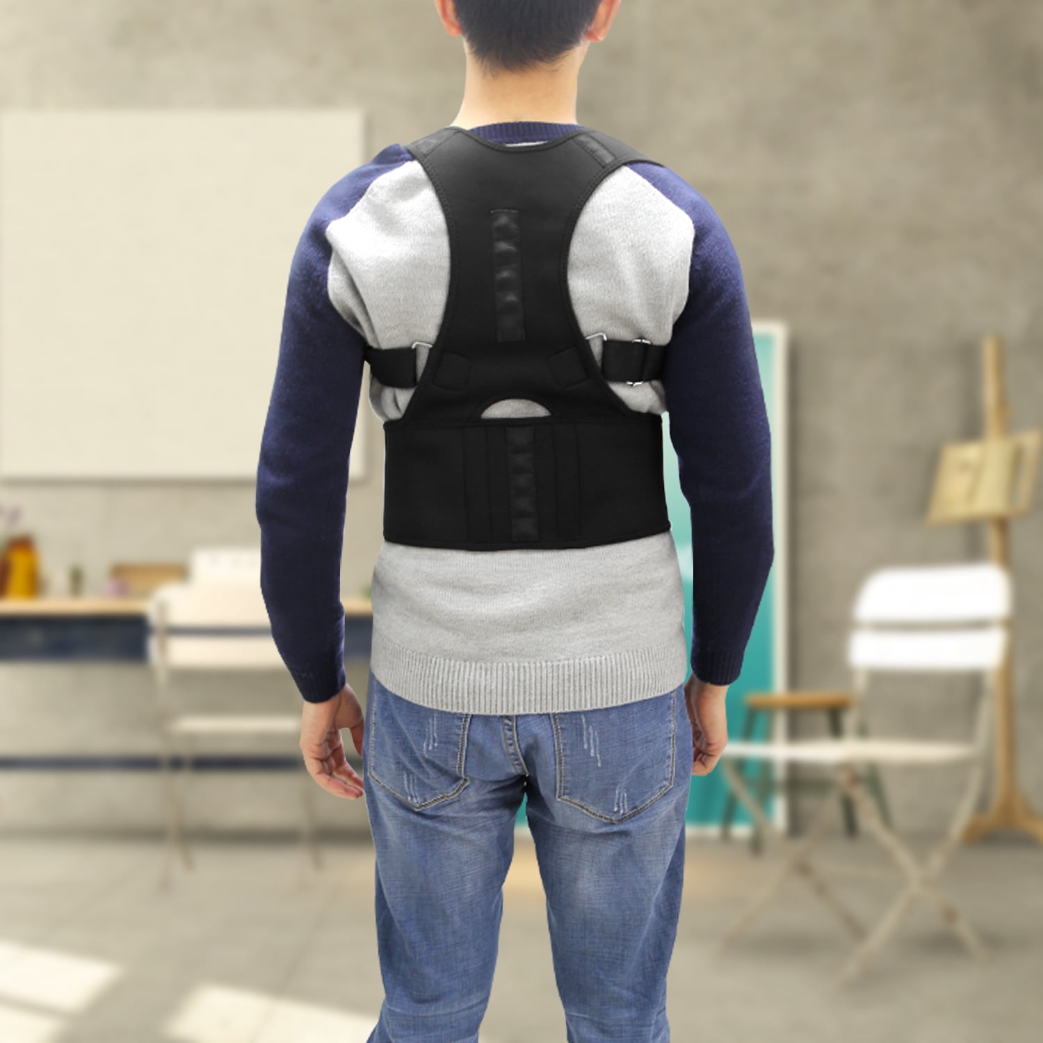 Eine Person, die eine Rückenstütze trägt, von hinten gesehen. Die Person trägt einen blauen Pullover, ein graues Hemd und Jeans, in einem Raum mit Möbeln.