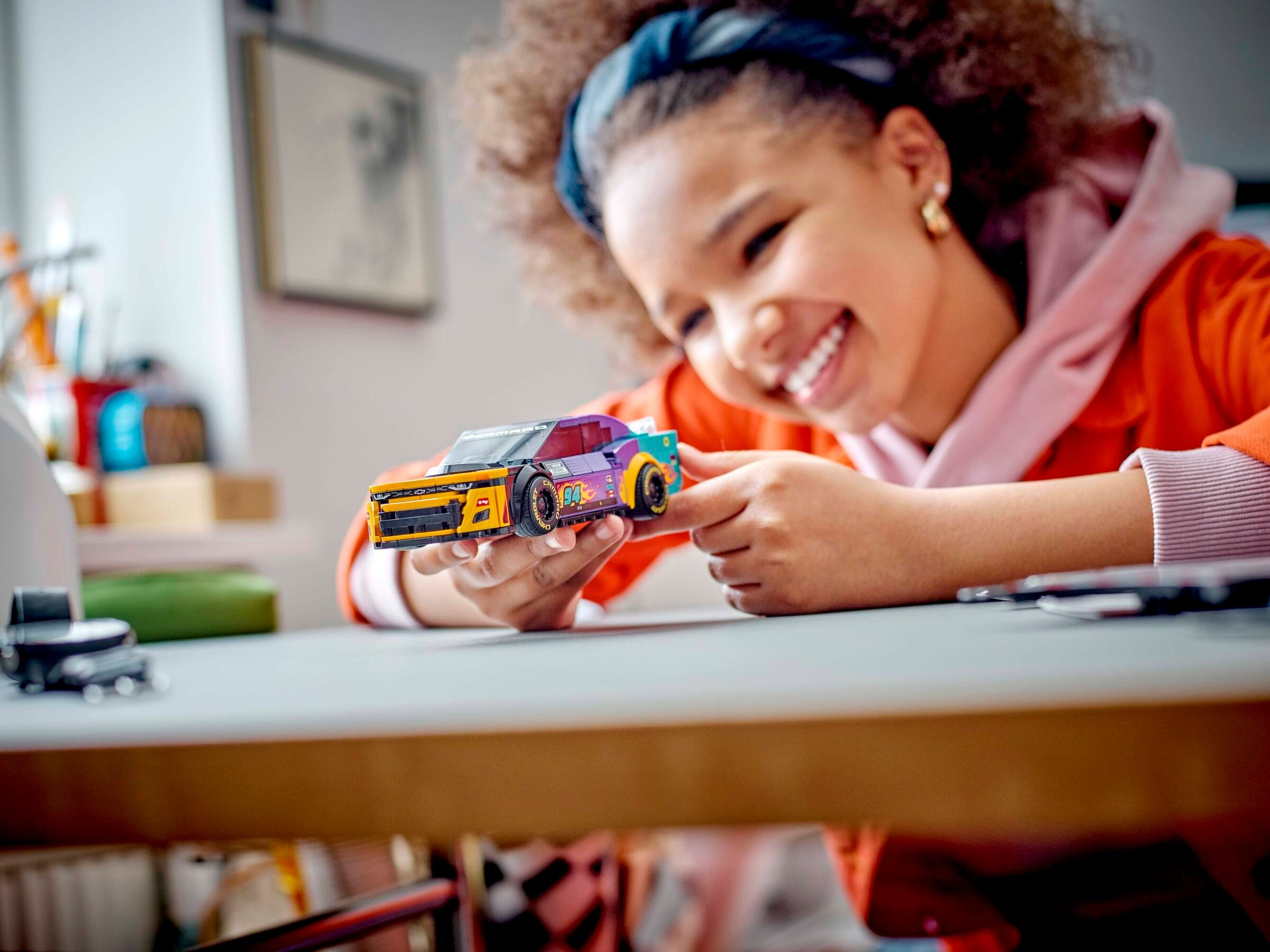 Ein lächelndes Mädchen hält ein Spielzeug-Rennwagen. Das Auto ist gelb, lila und blau. Sie trägt ein blaues Stirnband.