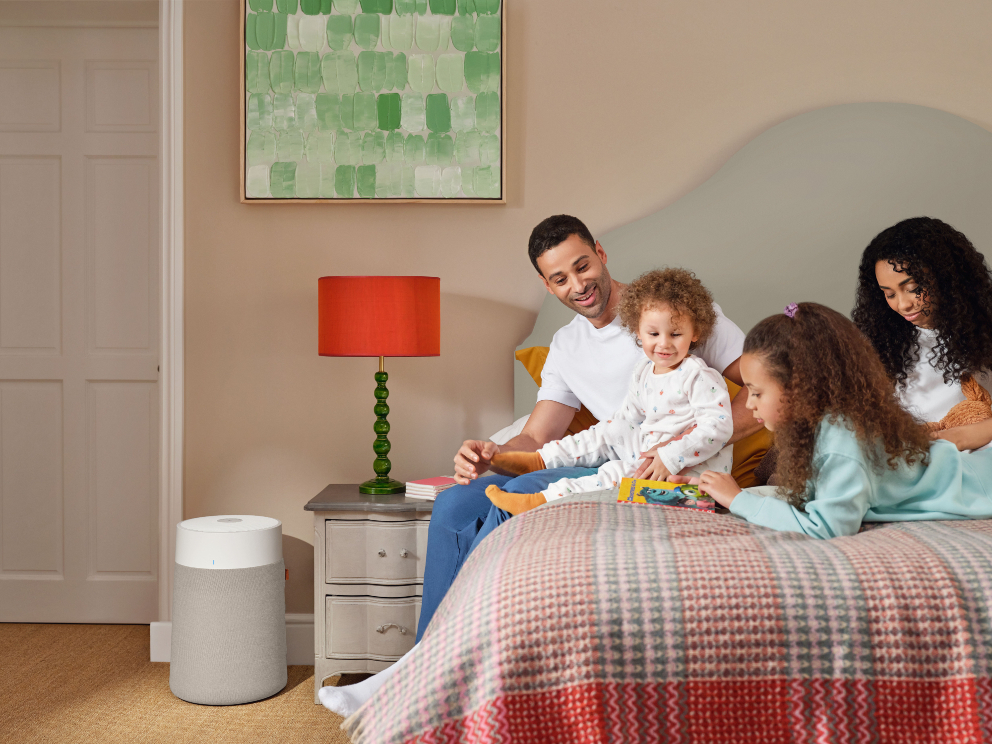 Una famiglia seduta su un letto. Un padre e due bambini leggono un libro. Sono visibili anche una lampada e un purificatore d'aria.