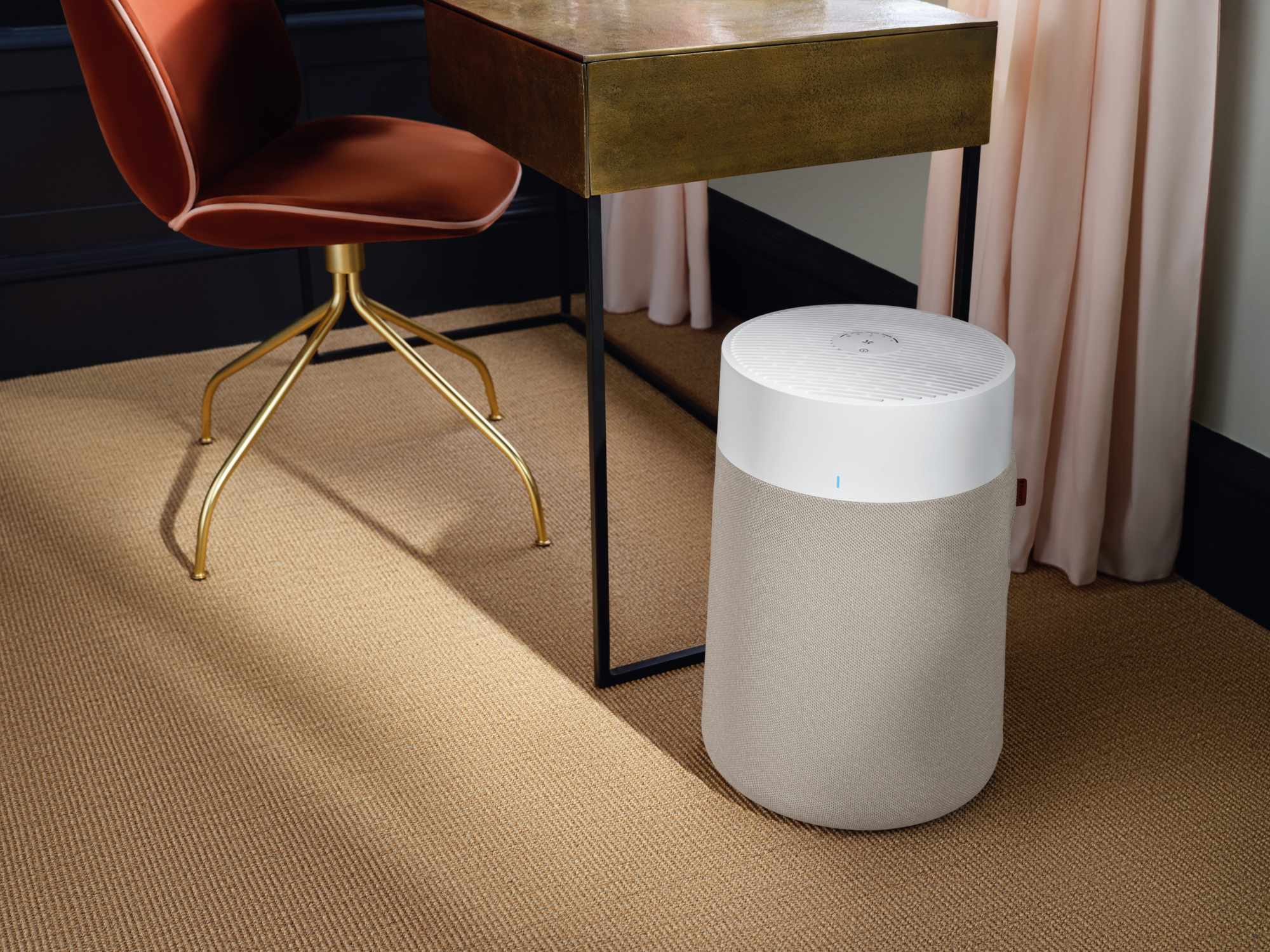 Un purificatore d'aria è accanto a una scrivania con una sedia marrone. La stanza ha una moquette beige e tende rosa.