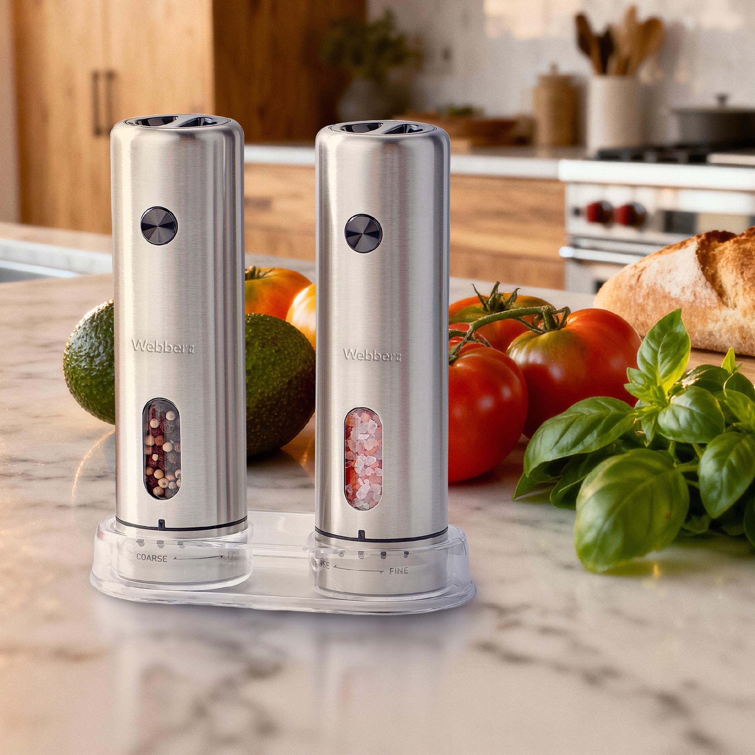 Dwa srebrne elektryczne młynki do soli i pieprzu na marmurowym blacie z awokado, pomidorami, chlebem i bazylią.
