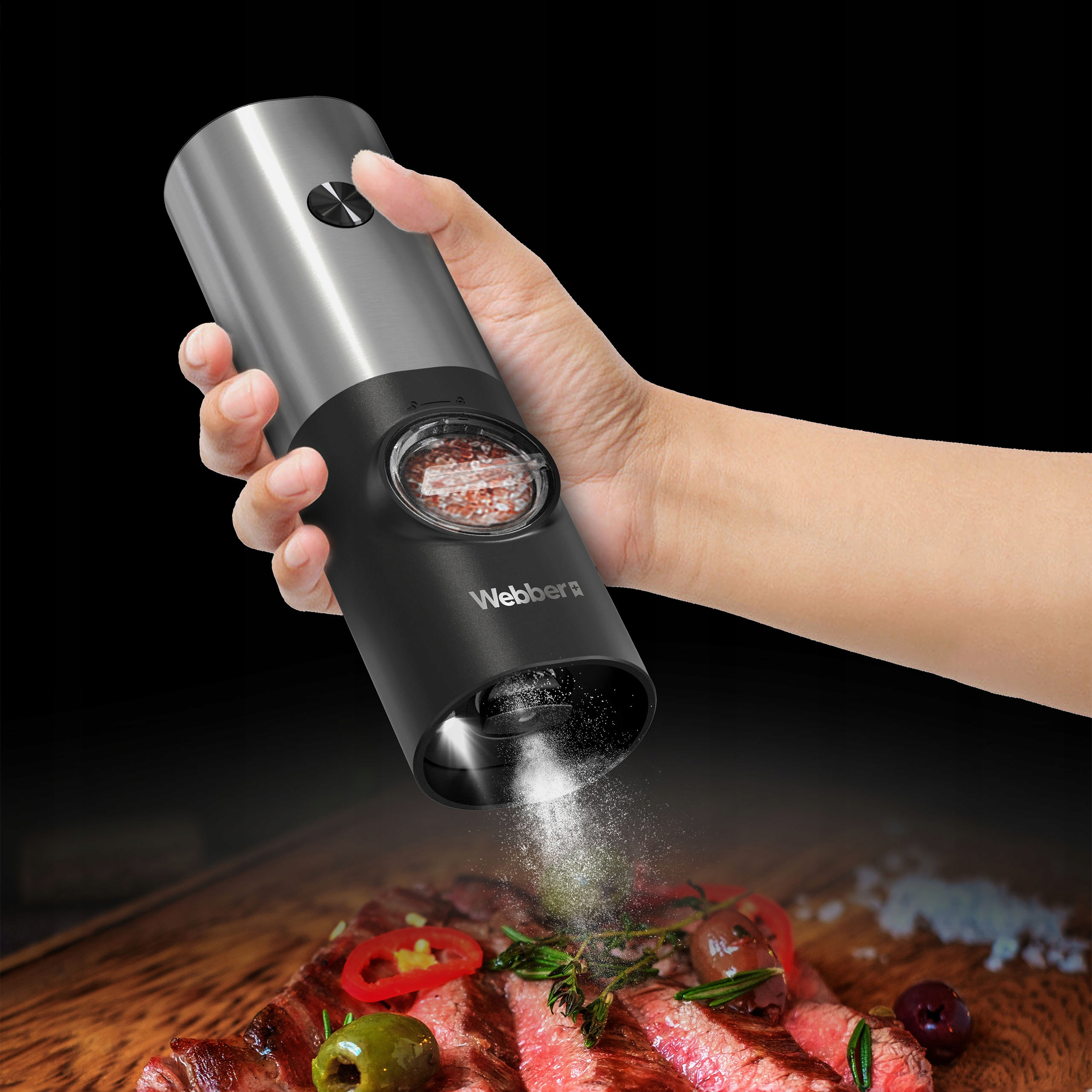Ręka trzyma młynek do soli Webber, posypując solą stek i warzywa na drewnianej desce.