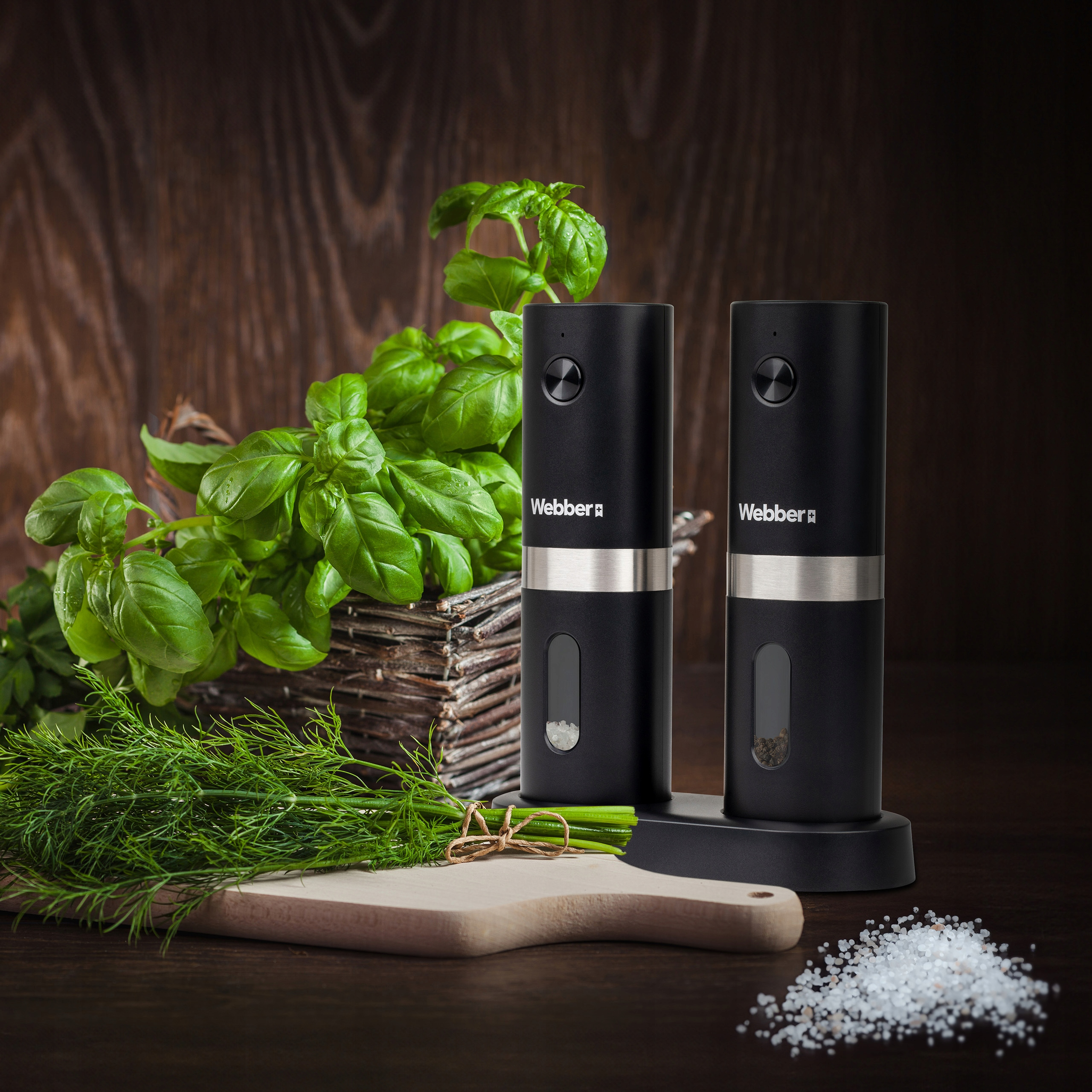 Dwa czarne młynki do soli i pieprzu, bazylia, koperek, drewniana deska do krojenia, sól i przyprawy na brązowym stole.