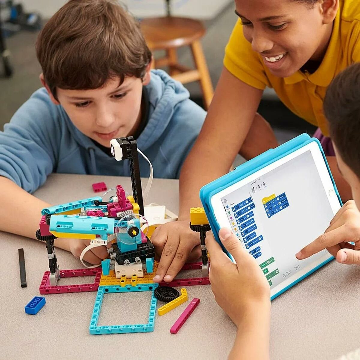 Tres niños construyen un robot con bloques coloridos. Uno sostiene una tableta con instrucciones de codificación, sonriendo. Los otros se enfocan en el robot.