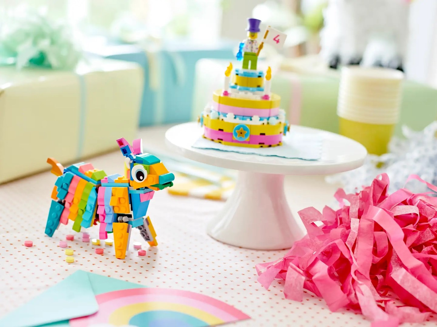 Kolorowa piniata Lego i ciasto na stole. Prezenty i różowe serpentyny je otaczają. Postać na cieście trzyma znak z numerem 4.