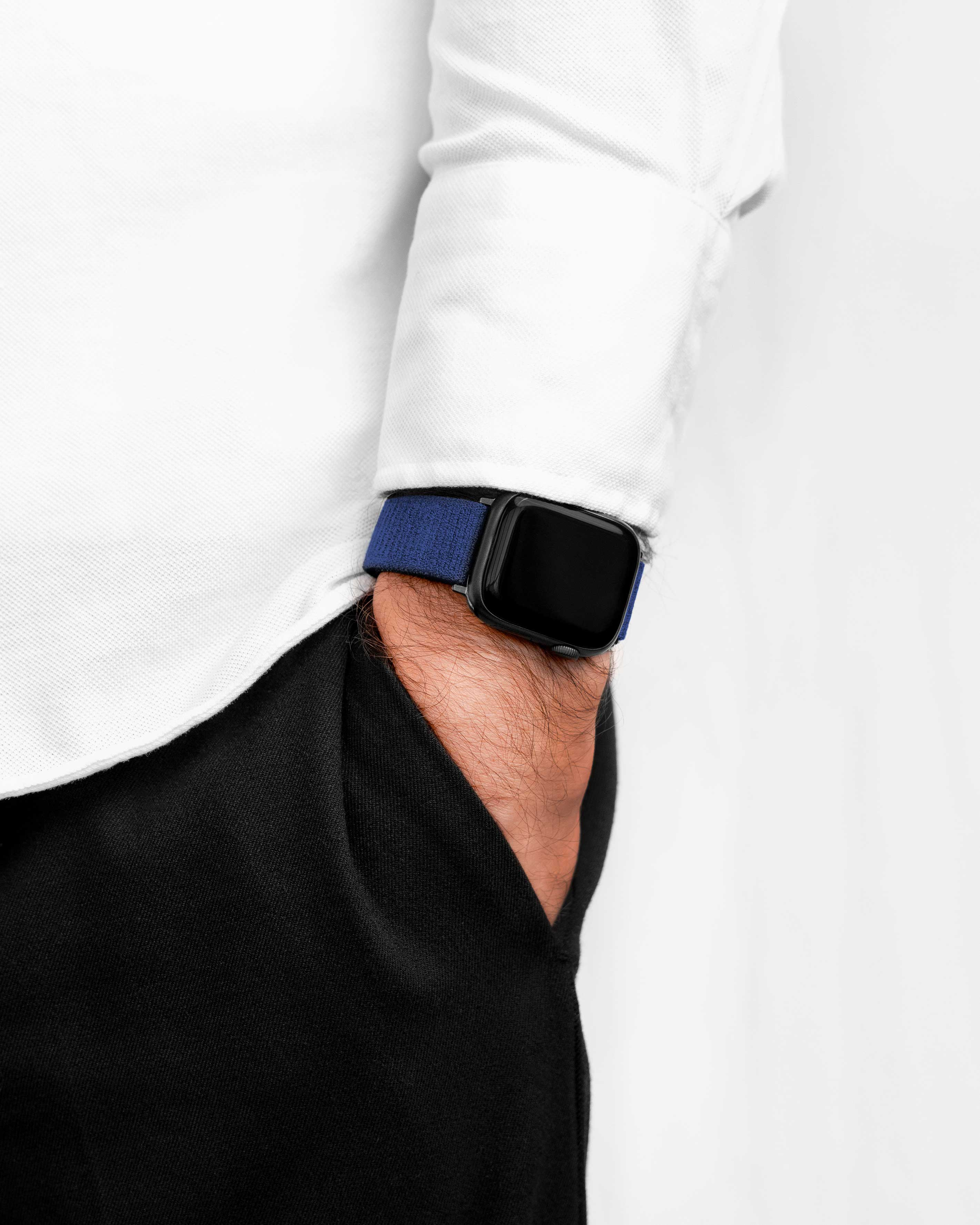 Arm einer Person mit blauem Uhrenarmband und schwarzer Uhr. Sie trägt ein weißes Hemd und schwarze Hose.