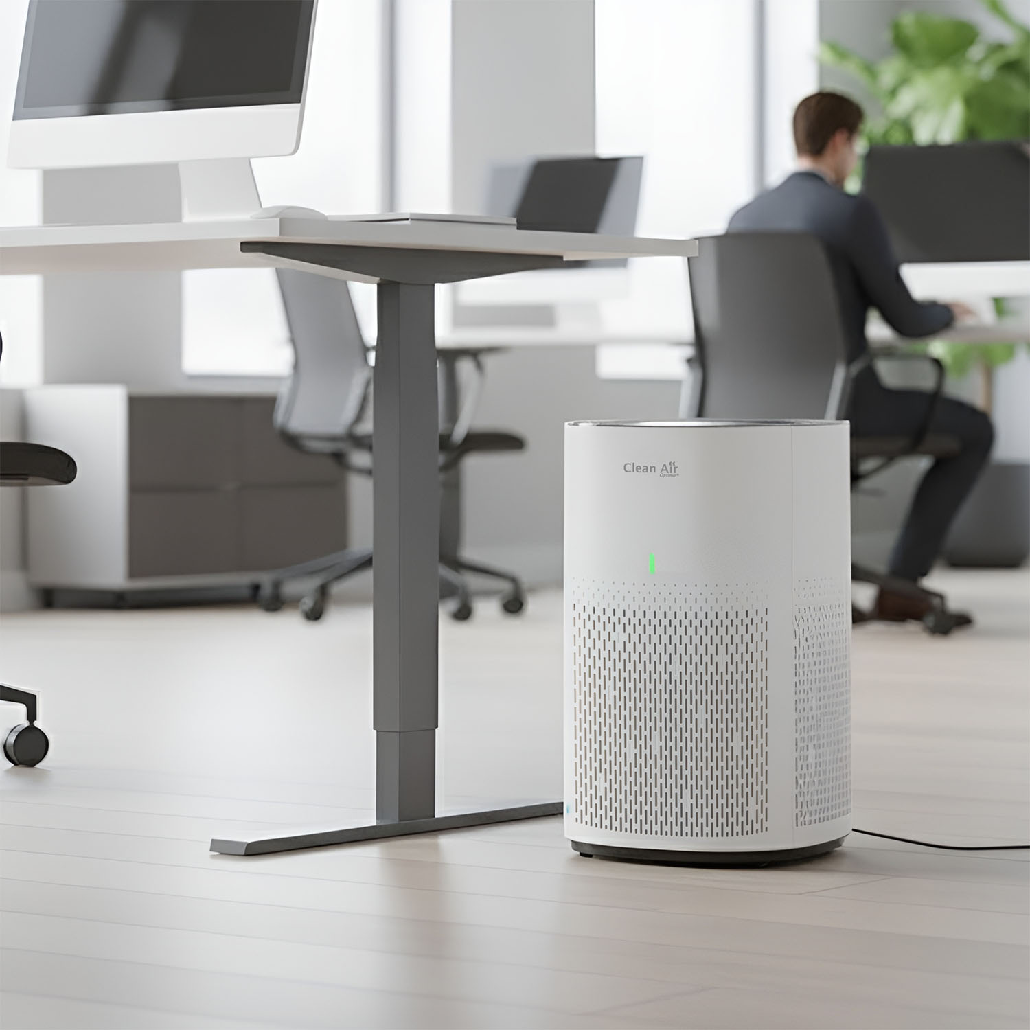 A white air purifier is on the floor in an office. A person is sitting at a desk. There are computers and desks in the background.