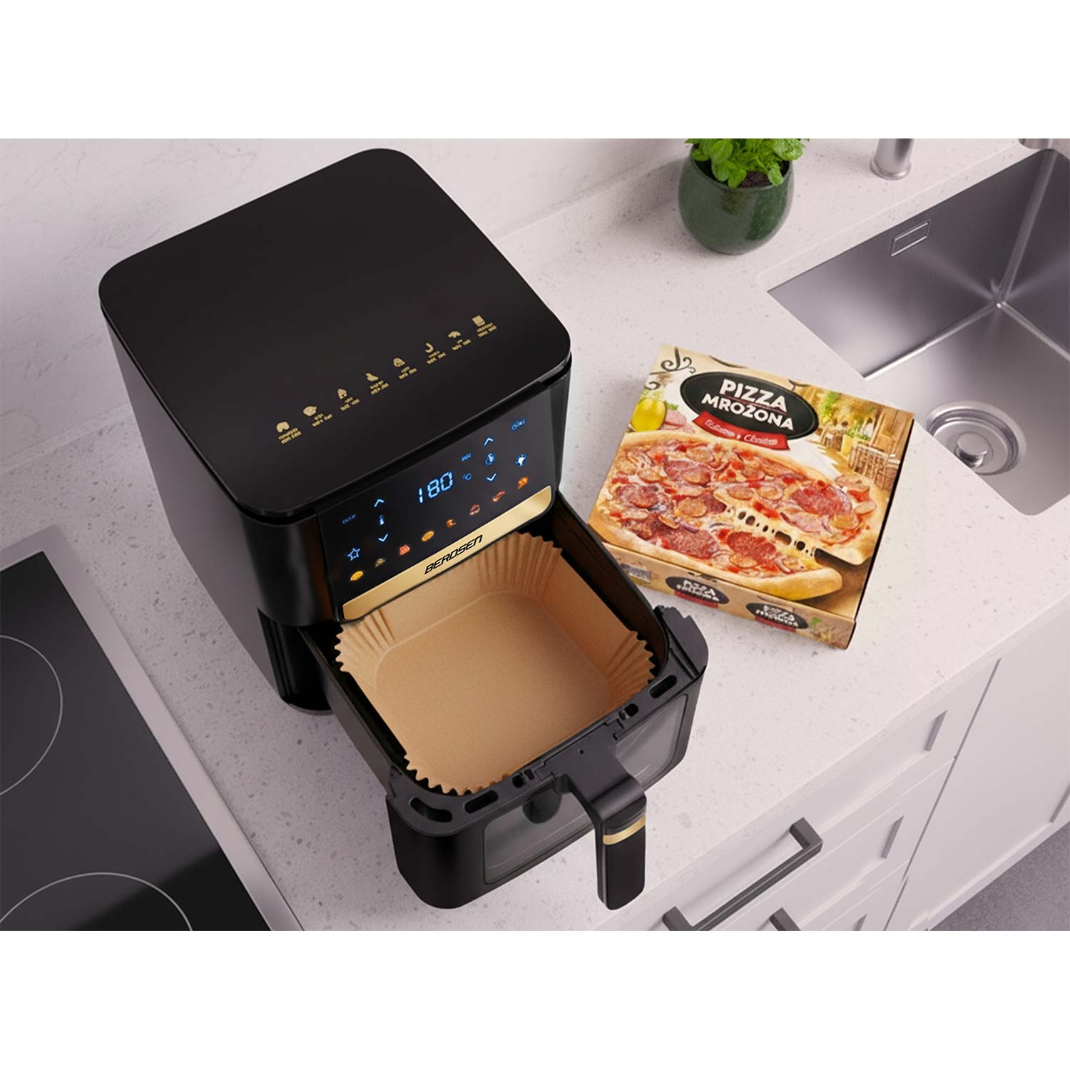 Czarna frytownica powietrzna z otwartą tacą i pudełkiem po pizzy w pobliżu na białym blacie. Roślina w tle.