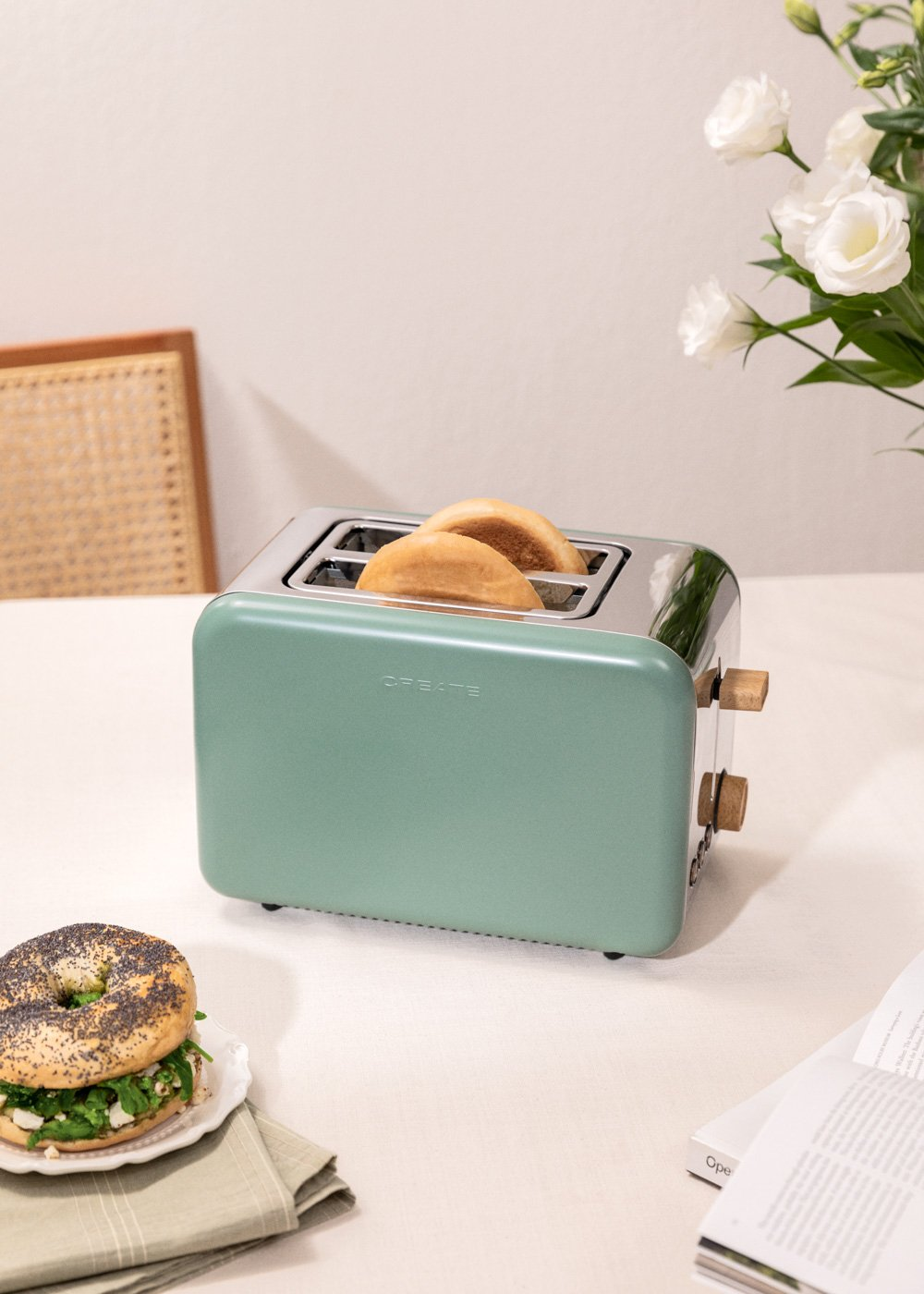 Una tostadora verde menta con pan tostado, un bagel y flores blancas sobre una mesa blanca.