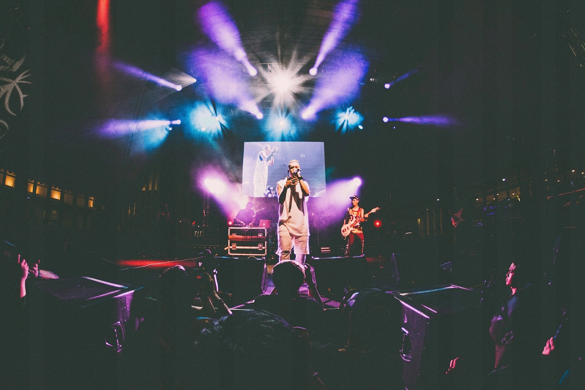 Piosenkarz na scenie, tłum na pierwszym planie. Scena oświetlona fioletowymi i białymi światłami. Gitarzysta po prawej.